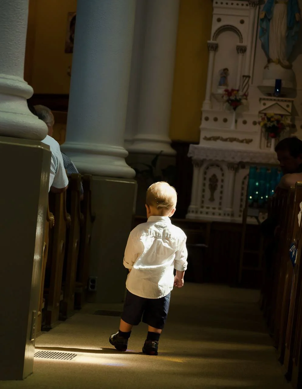 A young boy walking down the aisle of a church with sunlight illuminating his back.