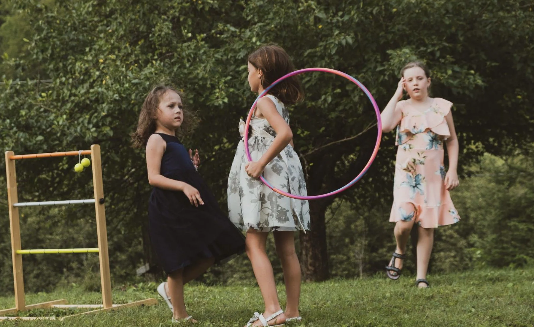 Three young girls playing in a park with a hula hoop and a wooden ladder with tennis balls hanging from it, surrounded by trees.