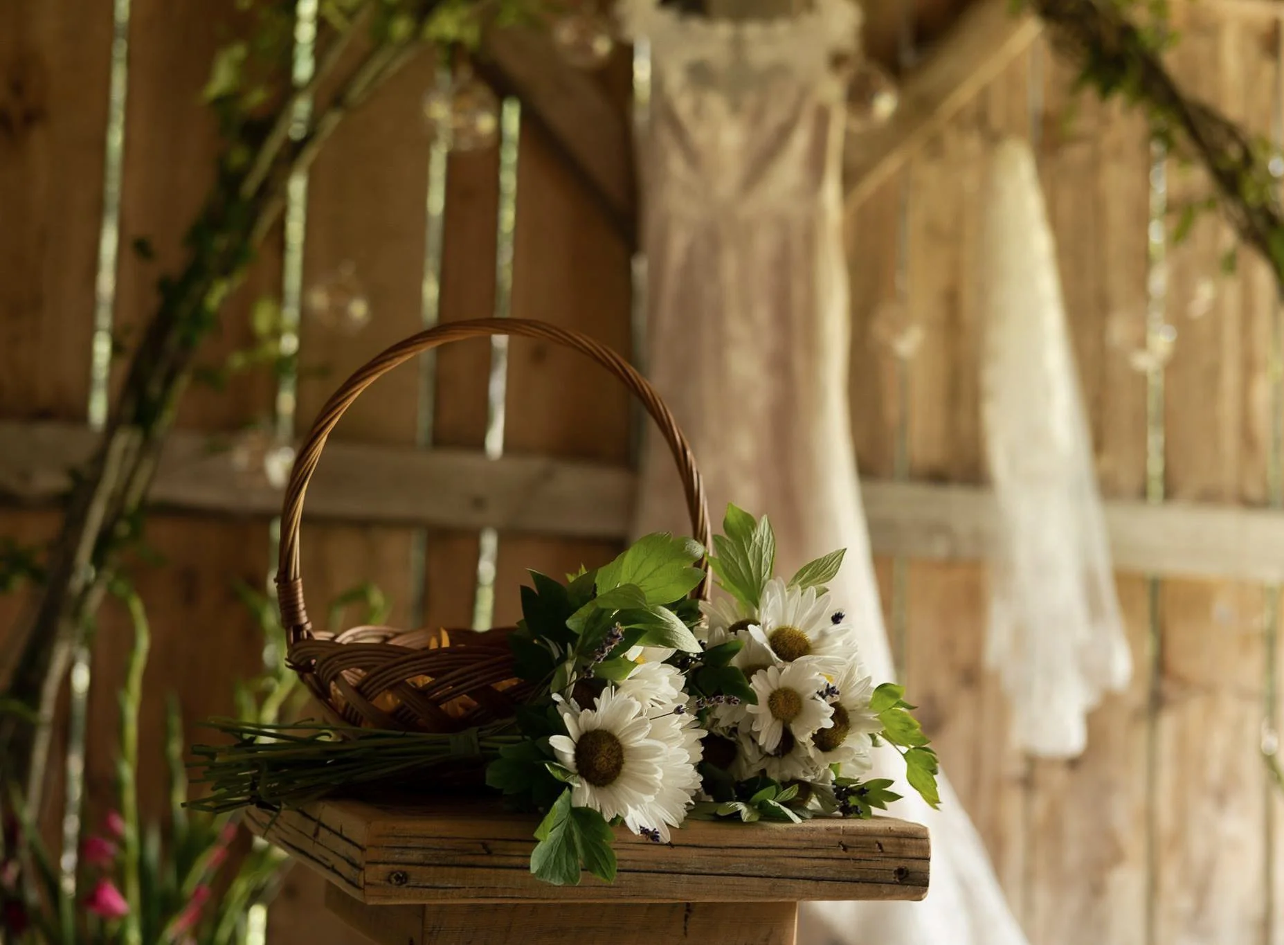 A woven basket filled with white daisies and green foliage, placed on a wooden surface in a rustic, outdoor setting with a wooden fence and trees in the background.