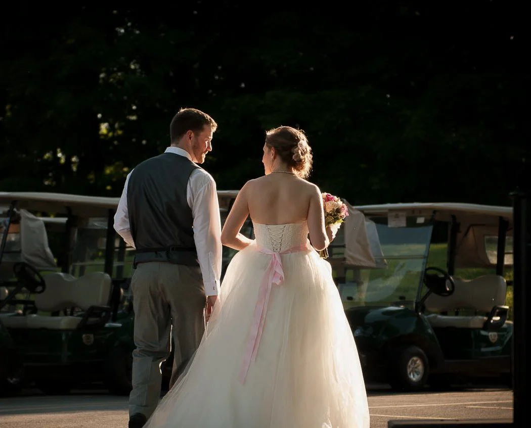 A bride and groom standing outside, facing each other. The bride is holding a bouquet, wearing a strapless wedding gown with a pink sash, and the groom is dressed in a white shirt and black vest. They are illuminated by sunlight, with golf carts and 