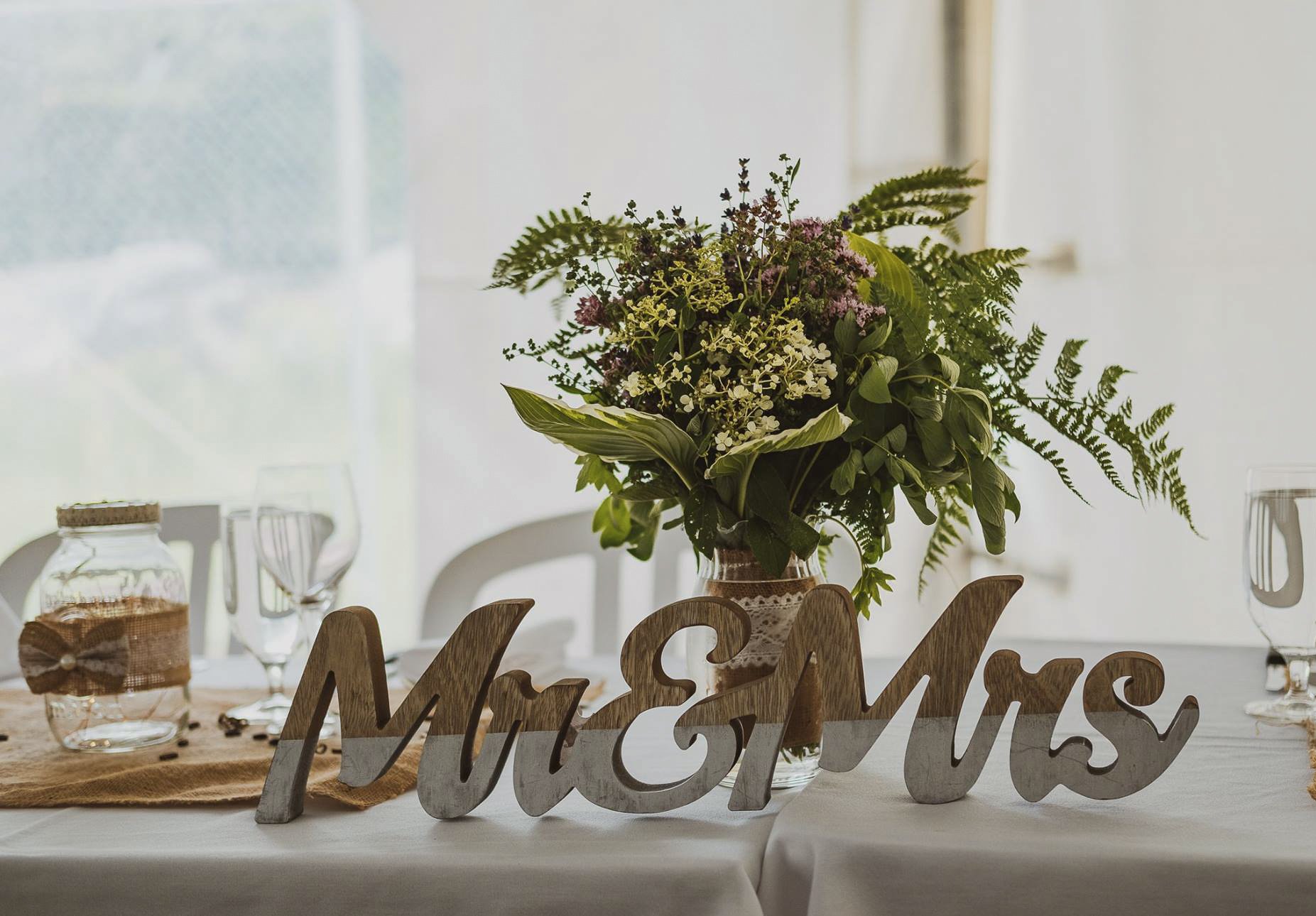 A floral centerpiece with greenery and small purple and white flowers on a dining table, with wooden chairs and a decorative 'Mr & Mrs' sign in the foreground.