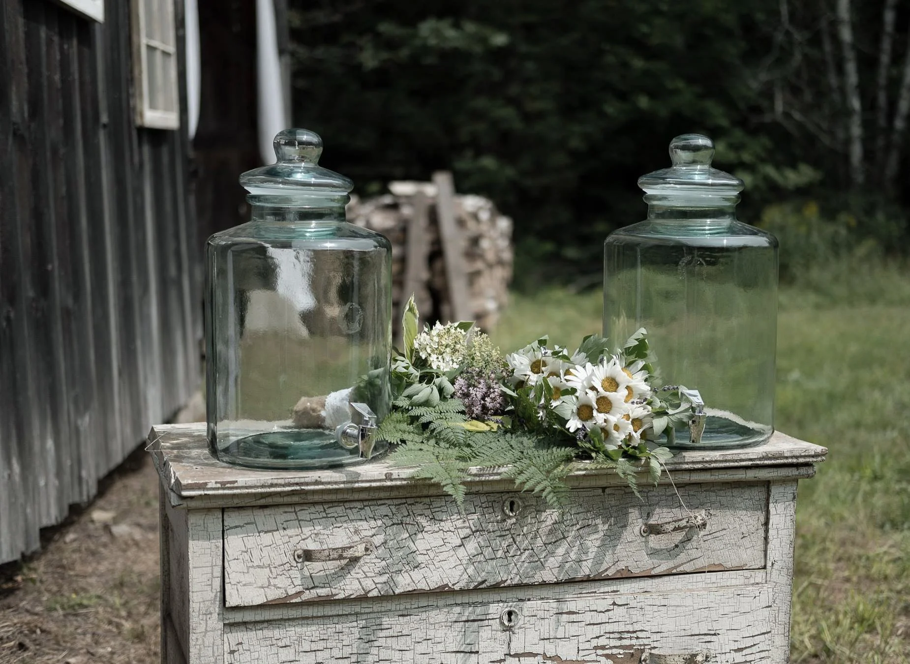 A rustic table with chipped white paint, holding two large glass apothecary jars with lids, surrounded by a floral arrangement of white daisies, purple flowers, and green foliage, outdoors near a weathered wooden building with a grassy background.
