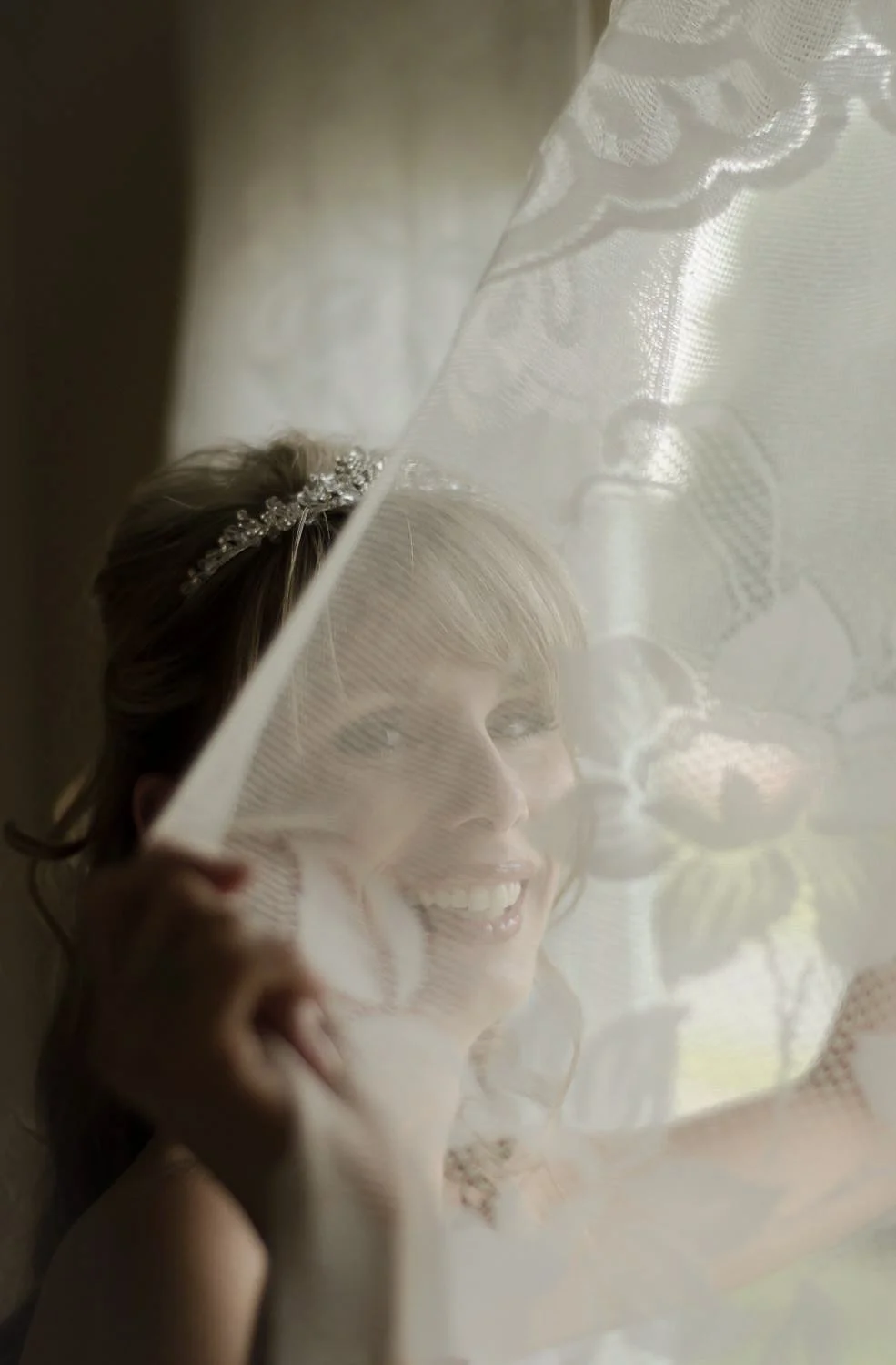 A bride with a bejeweled headband peeks through a sheer, embroidered curtain, smiling.