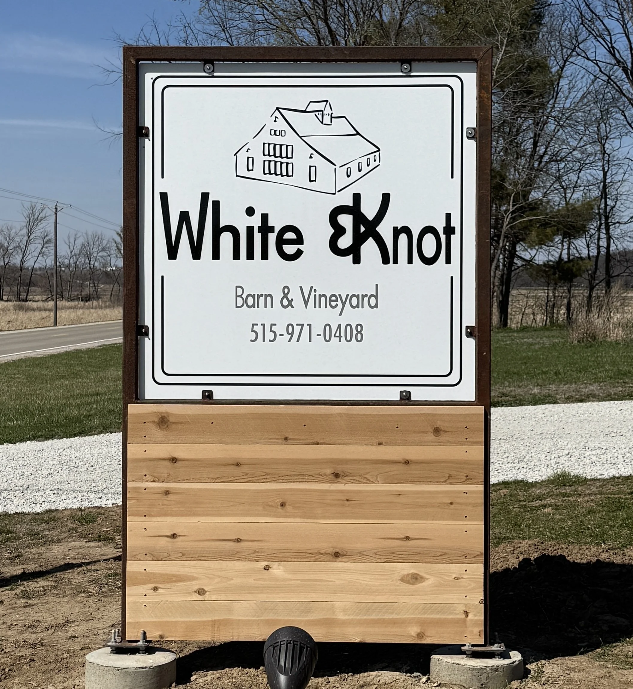 Sign for 'White & Knot' barn and vineyard with phone number 515-971-0408, featuring a simple house illustration, mounted on a wooden board with a landscape background.