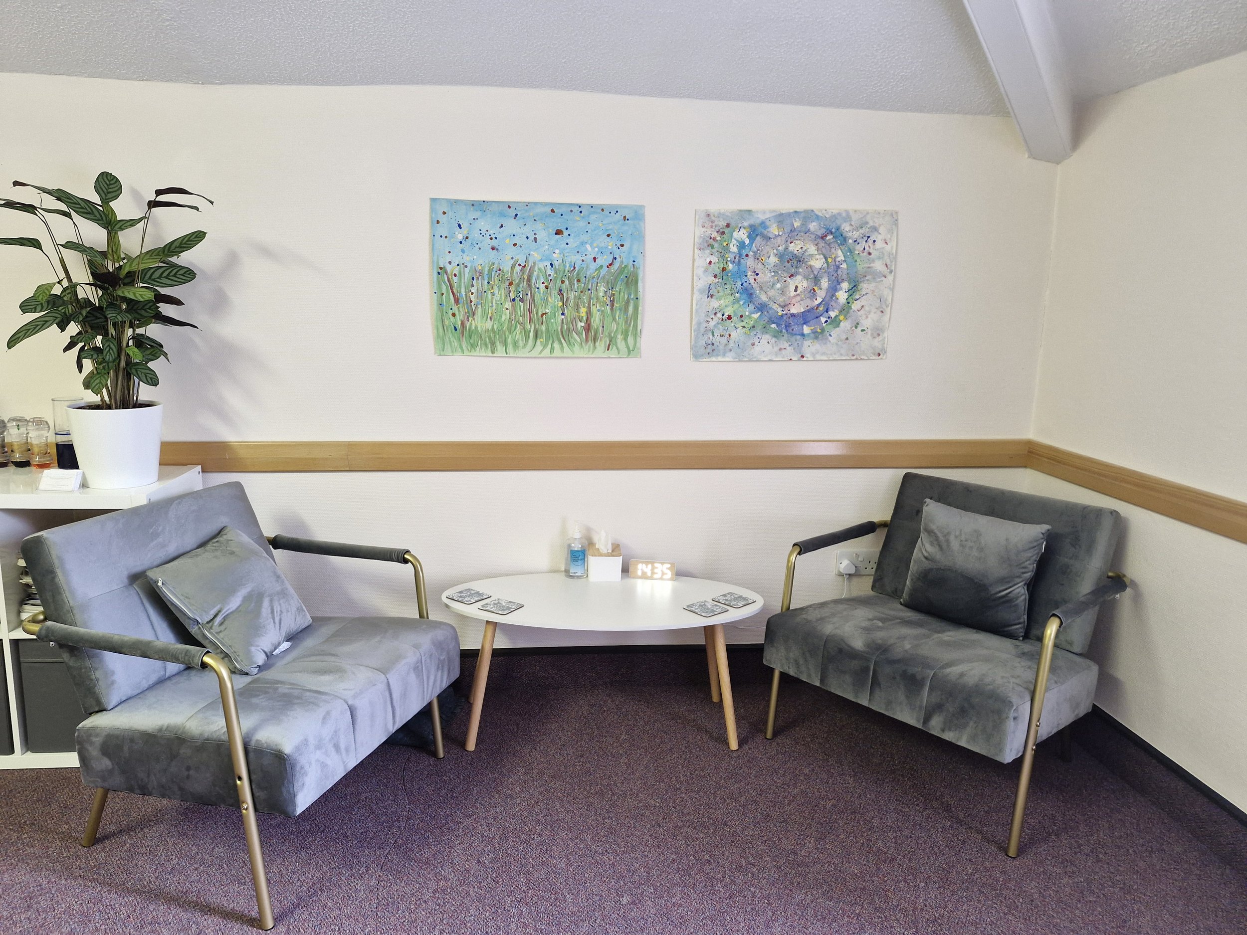 The image shows a counselling room with two chairs facing each other. There is a table between the chairs and some artwork on the wall behind the chairs.