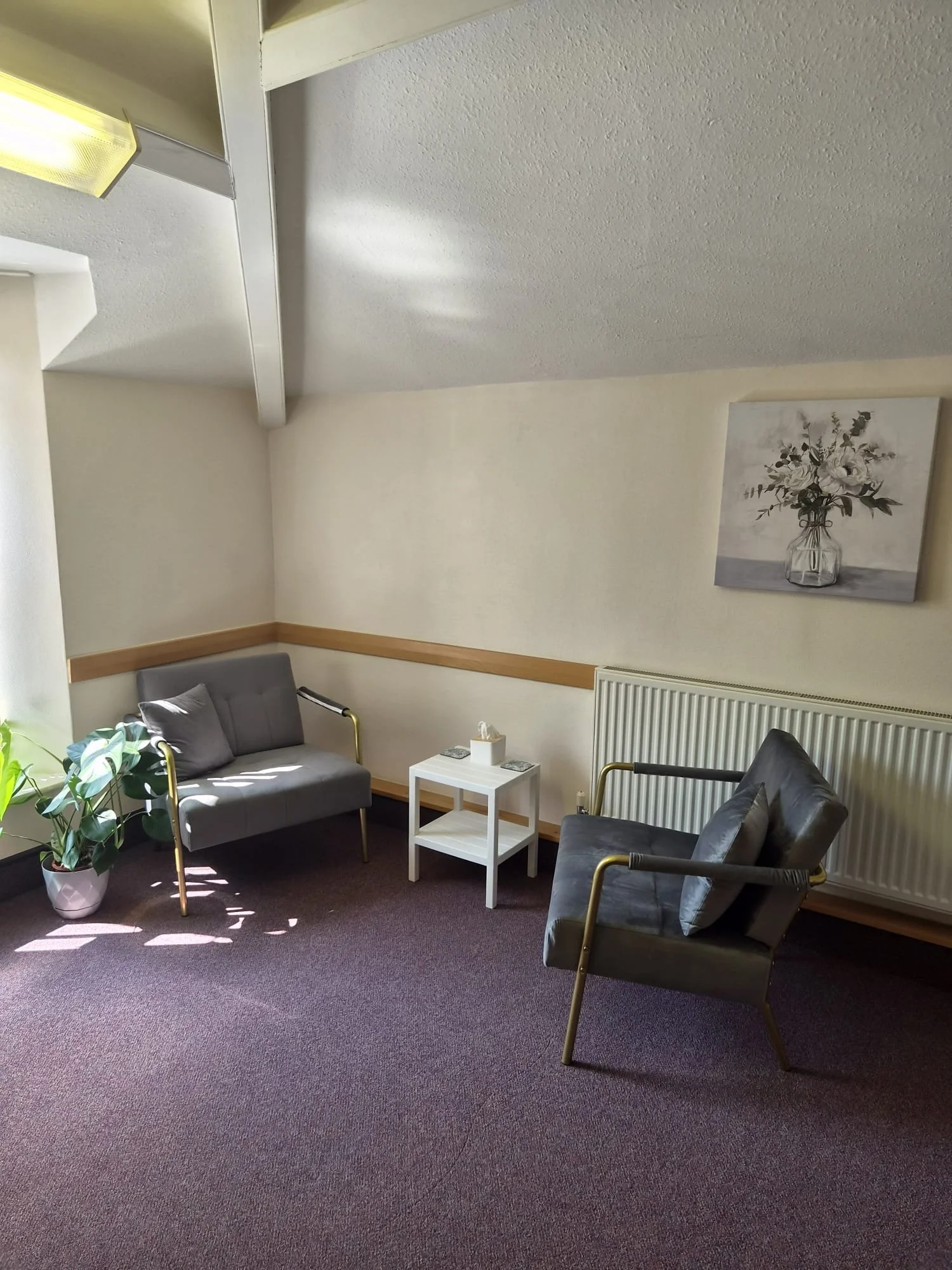 This image shows a counselling room with two chairs and a table. There is a canvas on the wall of flowers in a vase.