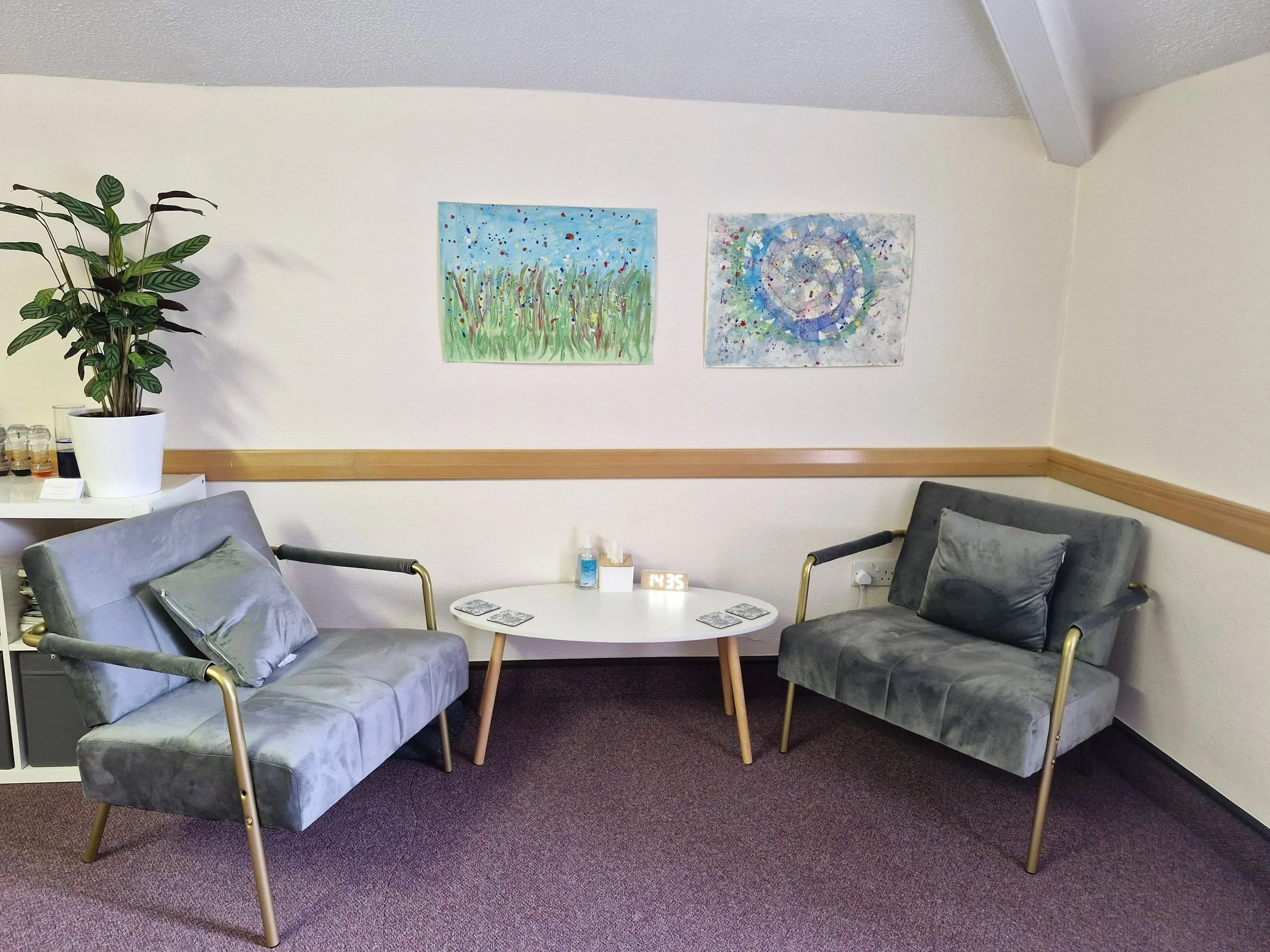 The image shows a counselling room with two grey armchair seats, and potted plant and some artwork on the wall. There is a table between the chairs with tissues and coasters.