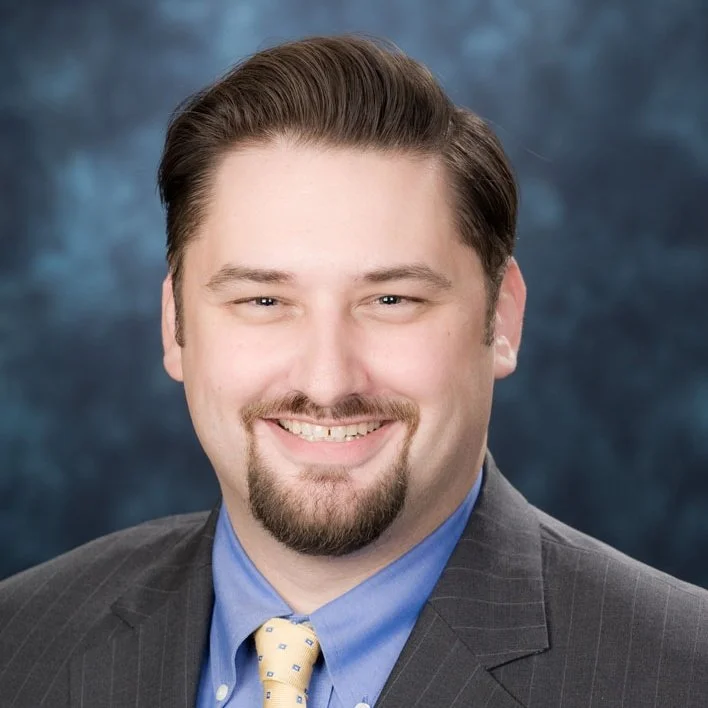 A professional headshot of a smiling man with short dark hair, a goatee, wearing a dark suit, blue dress shirt, and a yellow tie with small patterns, against a dark blue background.