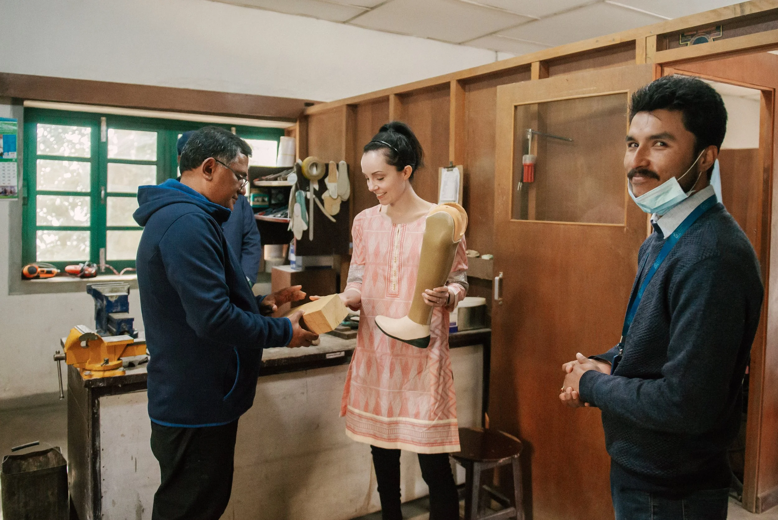 Stef holding a prosthetic leg while being shown a block of wood in a workshop.