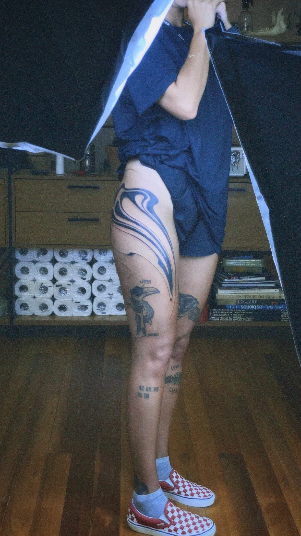 A person taking a mirror selfie, showing tattoos on their thigh and leg, wearing checkered slip-on shoes and standing in a room with shelves of books and toilet paper rolls.