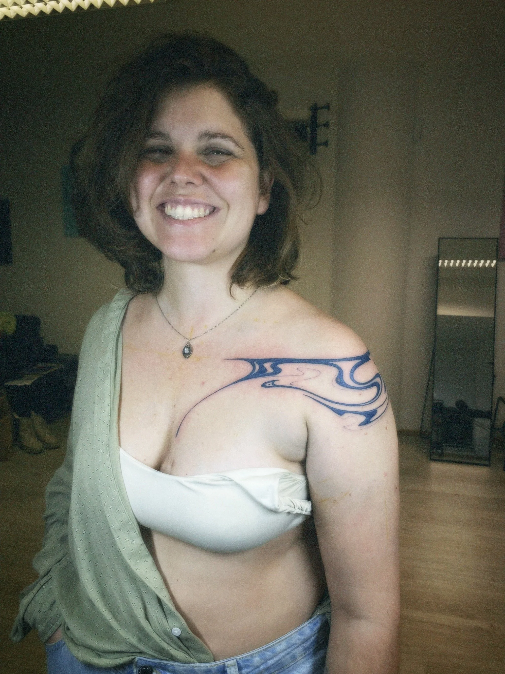 A smiling woman with shoulder-length brown hair, wearing a necklace, a falling-open light green shirt, a blue tattoo on her upper arm, and a white bra. She is standing in a room with a mirror, shelves, and shoes in the background.