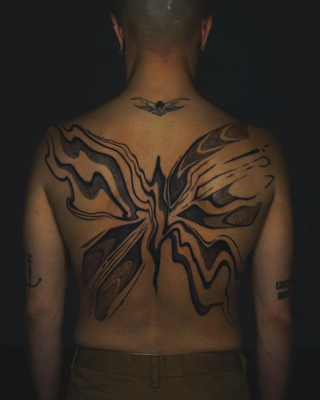 Person with a large abstract butterfly tattoo on their back and smaller tattoos on the arms.