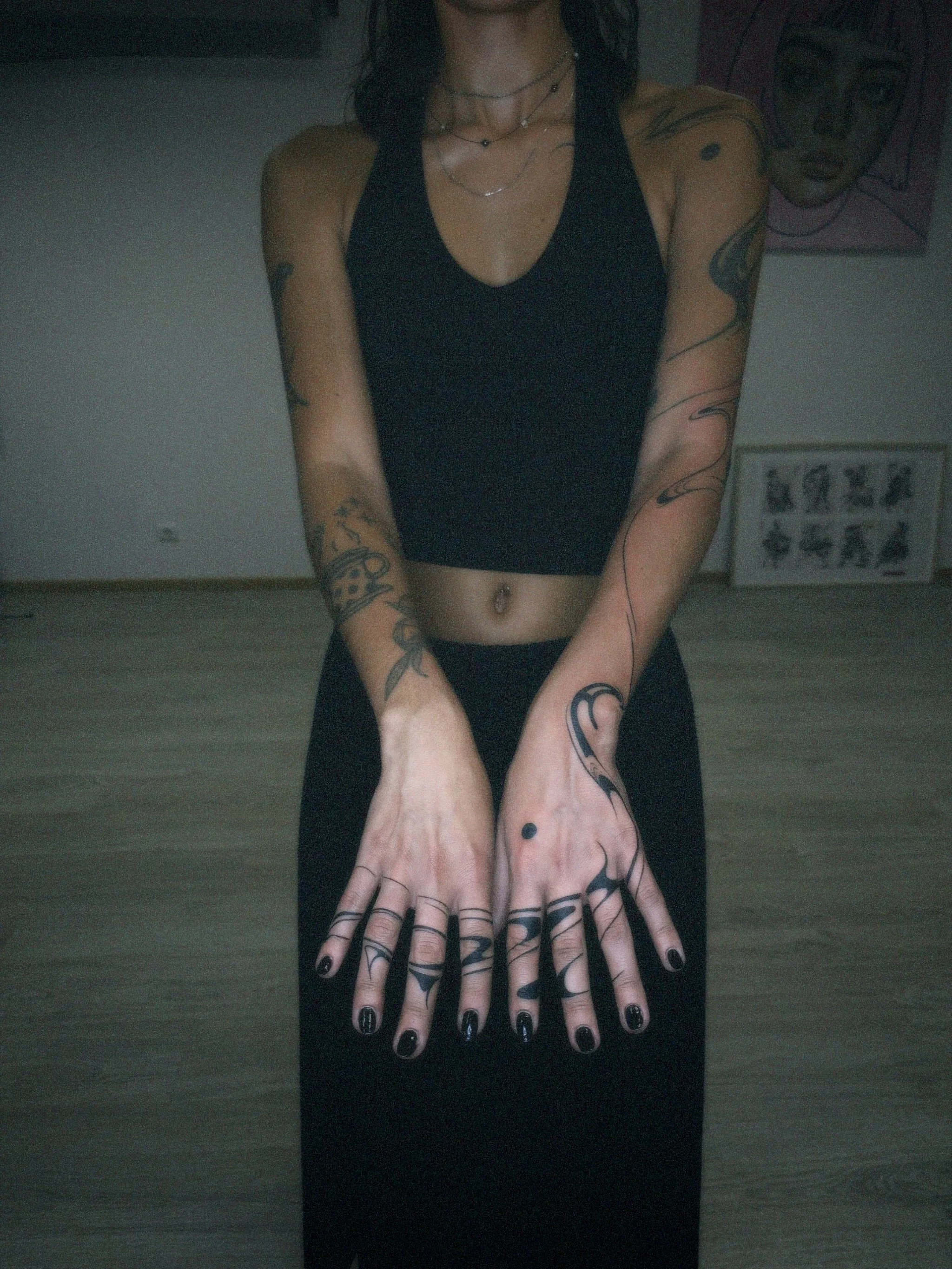 A woman with tattoos on her arms and torso displaying her hands with black nail polish and rings, standing in a room with artwork on the walls.