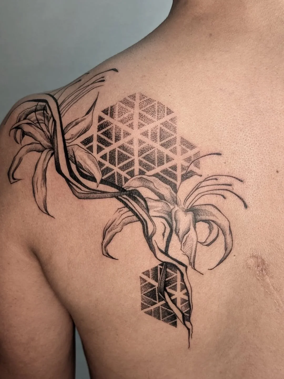 A black and grey tattoo of a lily flower with geometric triangles on the person's shoulder and upper chest.