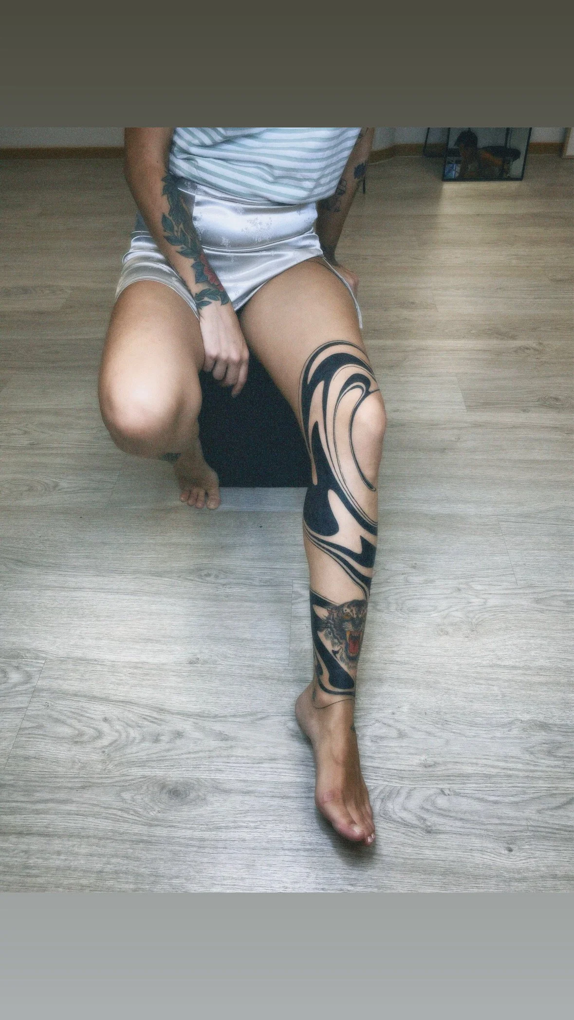Person sitting on a black stool with tattoos on their left arm and a large black and gray tattoo on their right leg, wearing a striped shirt and shiny silver shorts, bare feet on a light wooden floor, with a small dog in a mirror in the background.