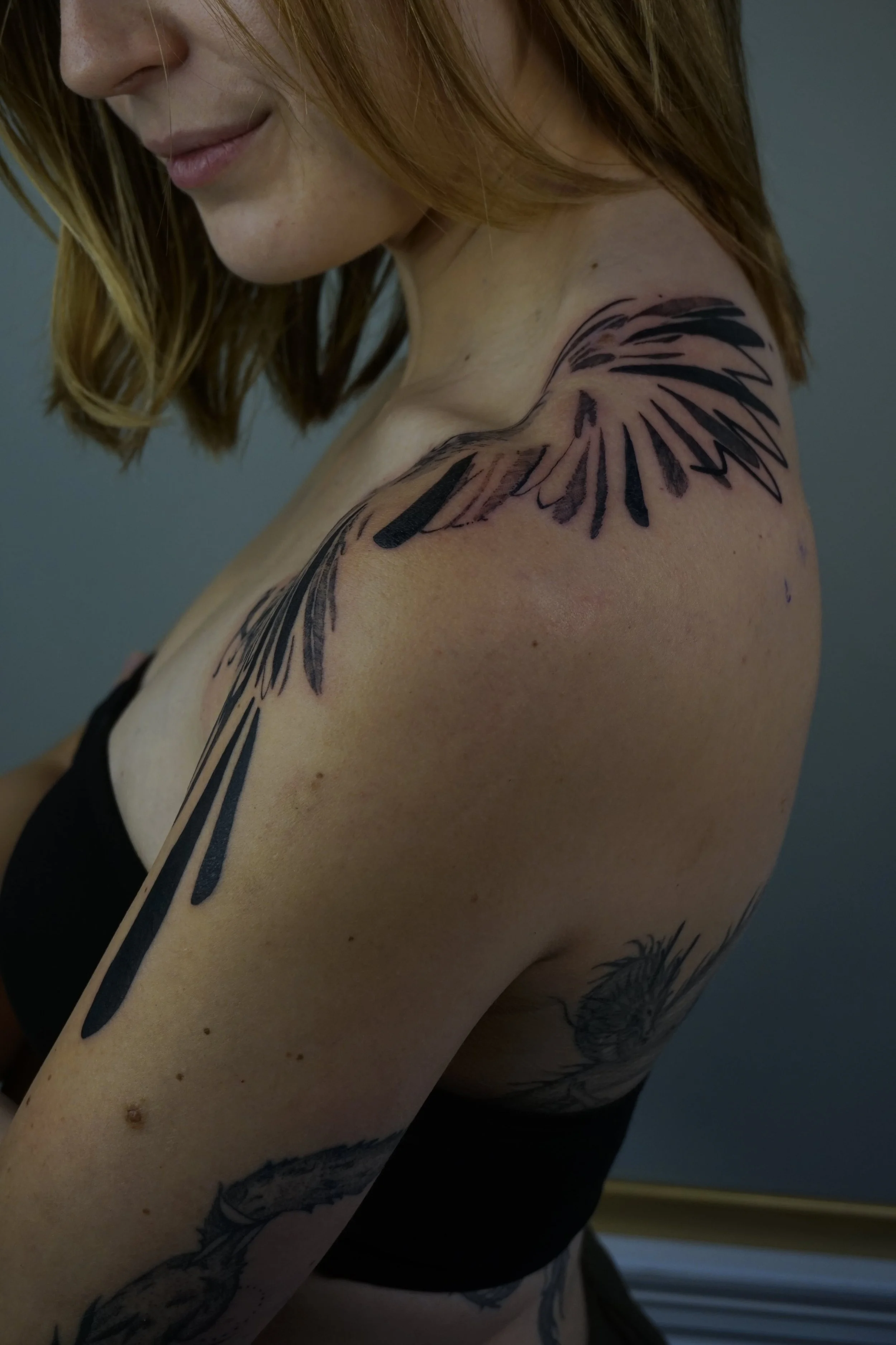 A woman with long red hair shows a partially completed tattoo of feathers or wings on her shoulder and upper arm.