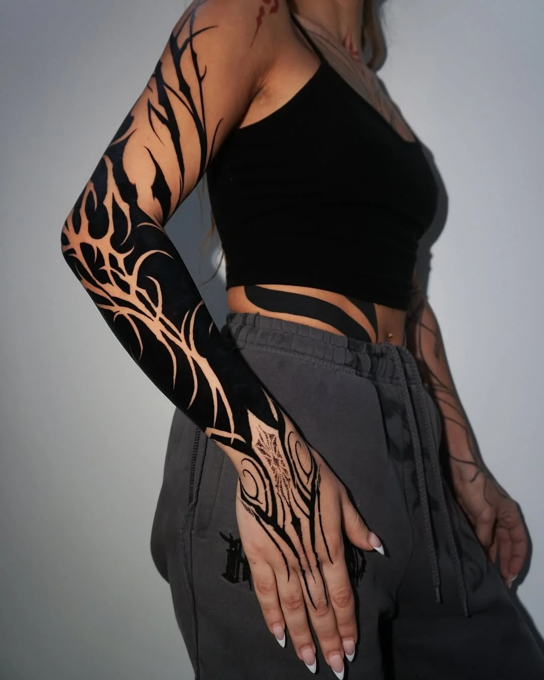 A woman with tattoos on her arms and side wearing a black crop top and gray jogger pants.