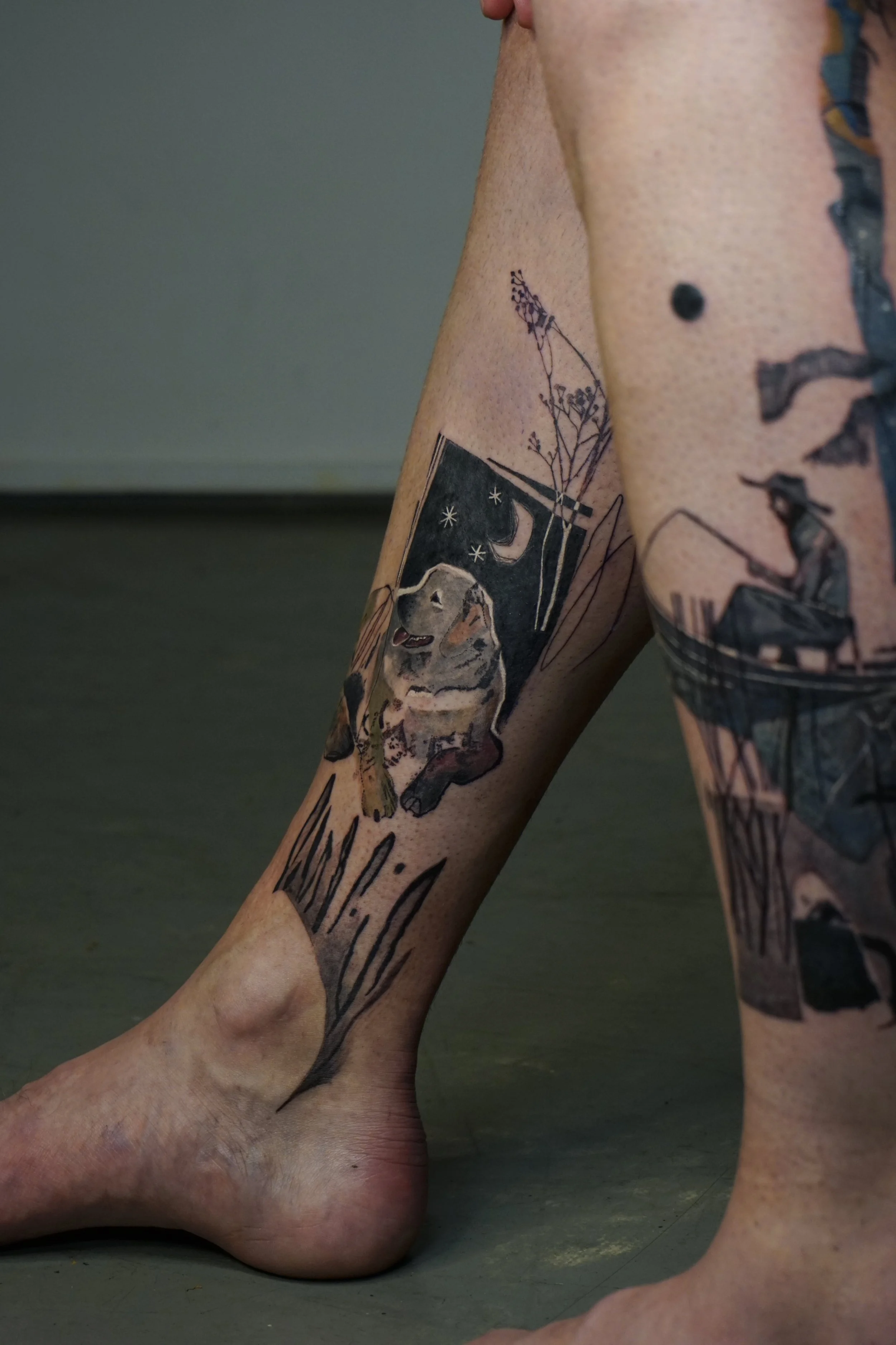 Close-up of a person's lower leg with multiple tattoos, including a dog, a moon, stars, and floral designs.