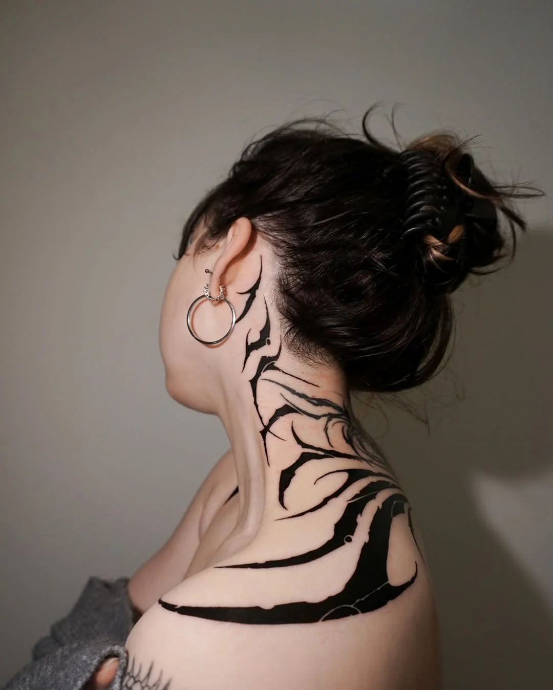 A woman with dark hair styled in a messy bun wearing large hoop earrings has a black and white tribal tattoo on her neck and shoulder, and is turned sideways against a neutral background.