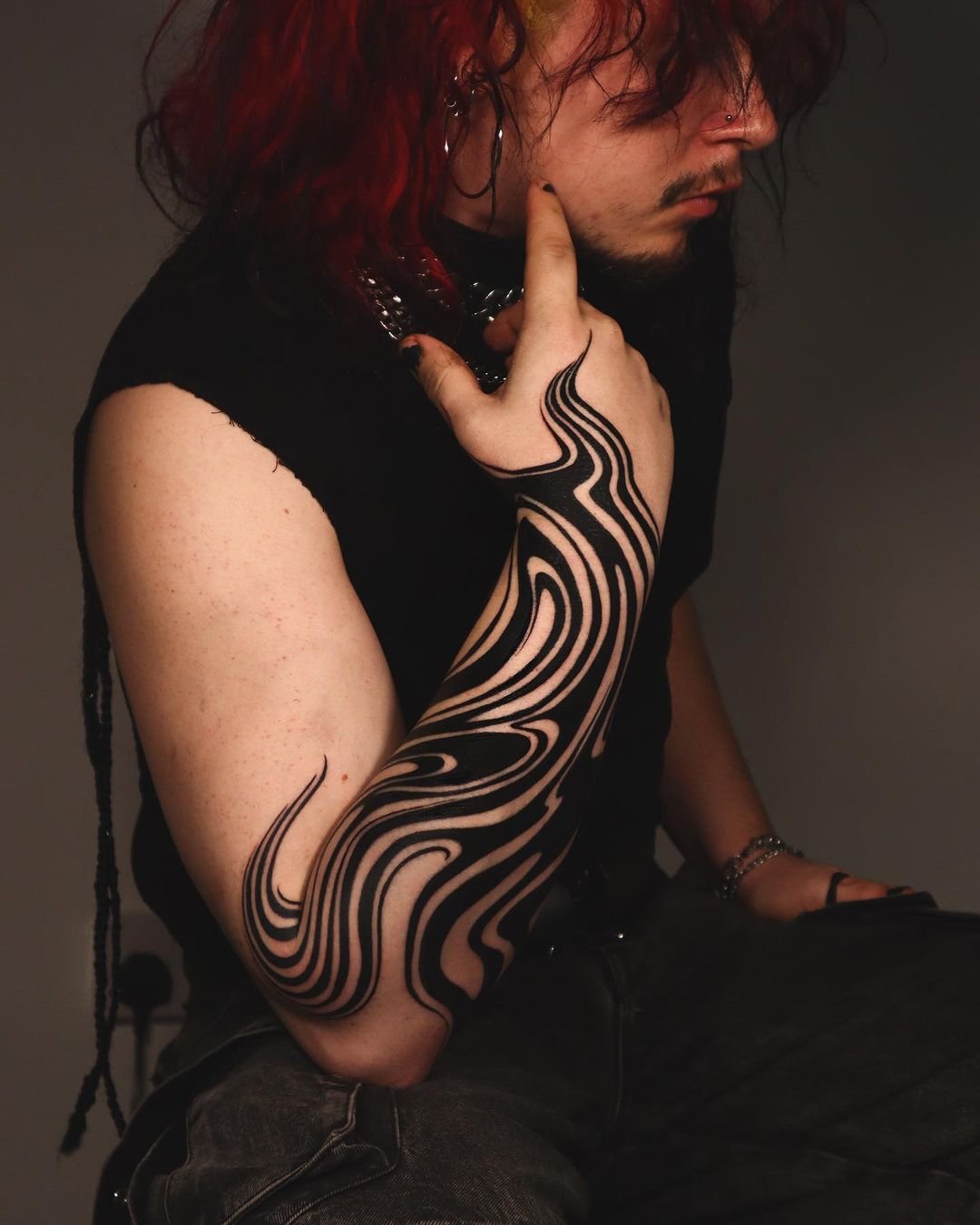 Person with red hair and intricate black arm tattoo, wearing a sleeveless shirt and silver bracelet, touching their chin.