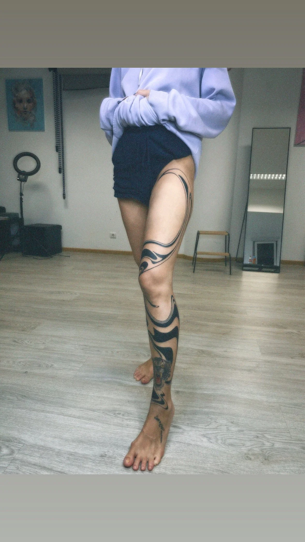 A person standing in a room with wooden flooring, wearing dark shorts and a light-colored hoodie, with visible tattoos on their leg.