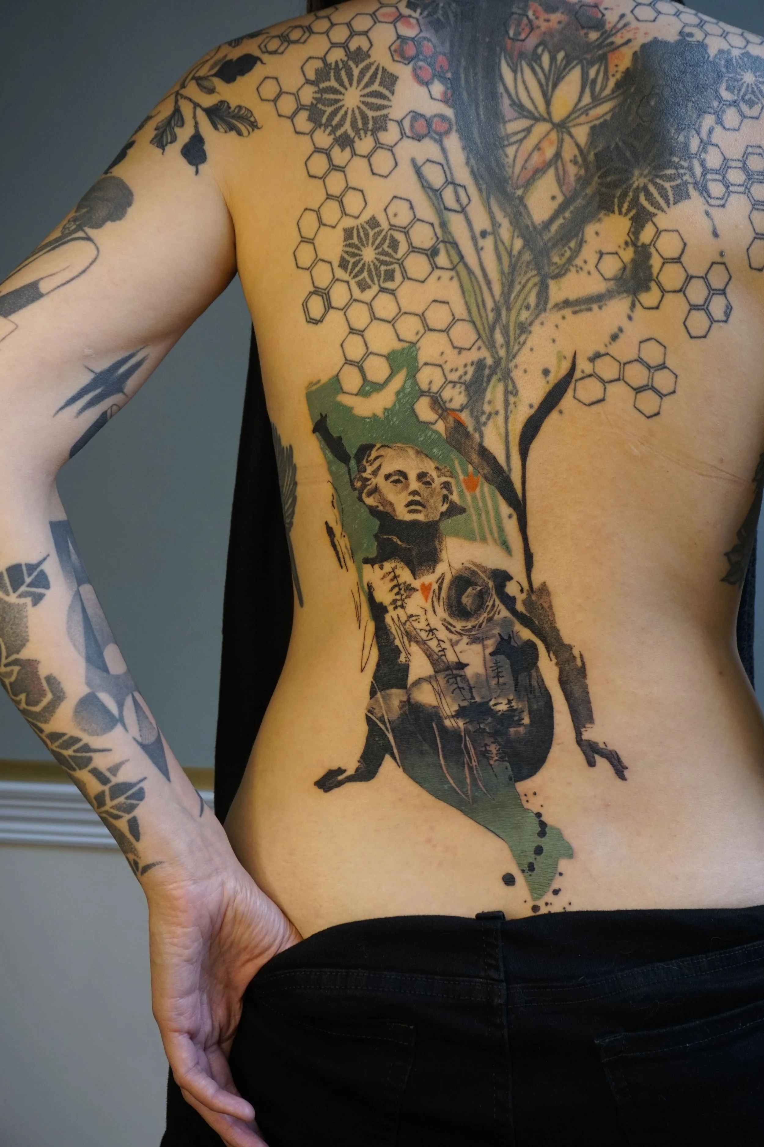 A person with tattoos on their chest and arms, featuring floral, honeycomb, and geometric patterns, along with a large tattoo of a woman sitting down, mixed with abstract and nature elements.
