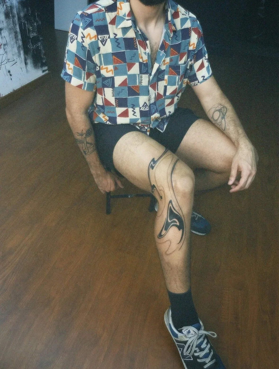 Person sitting on a chair, wearing a colorful patterned short-sleeve shirt, black shorts, and sneakers. They have tattoos on both arms and their legs.