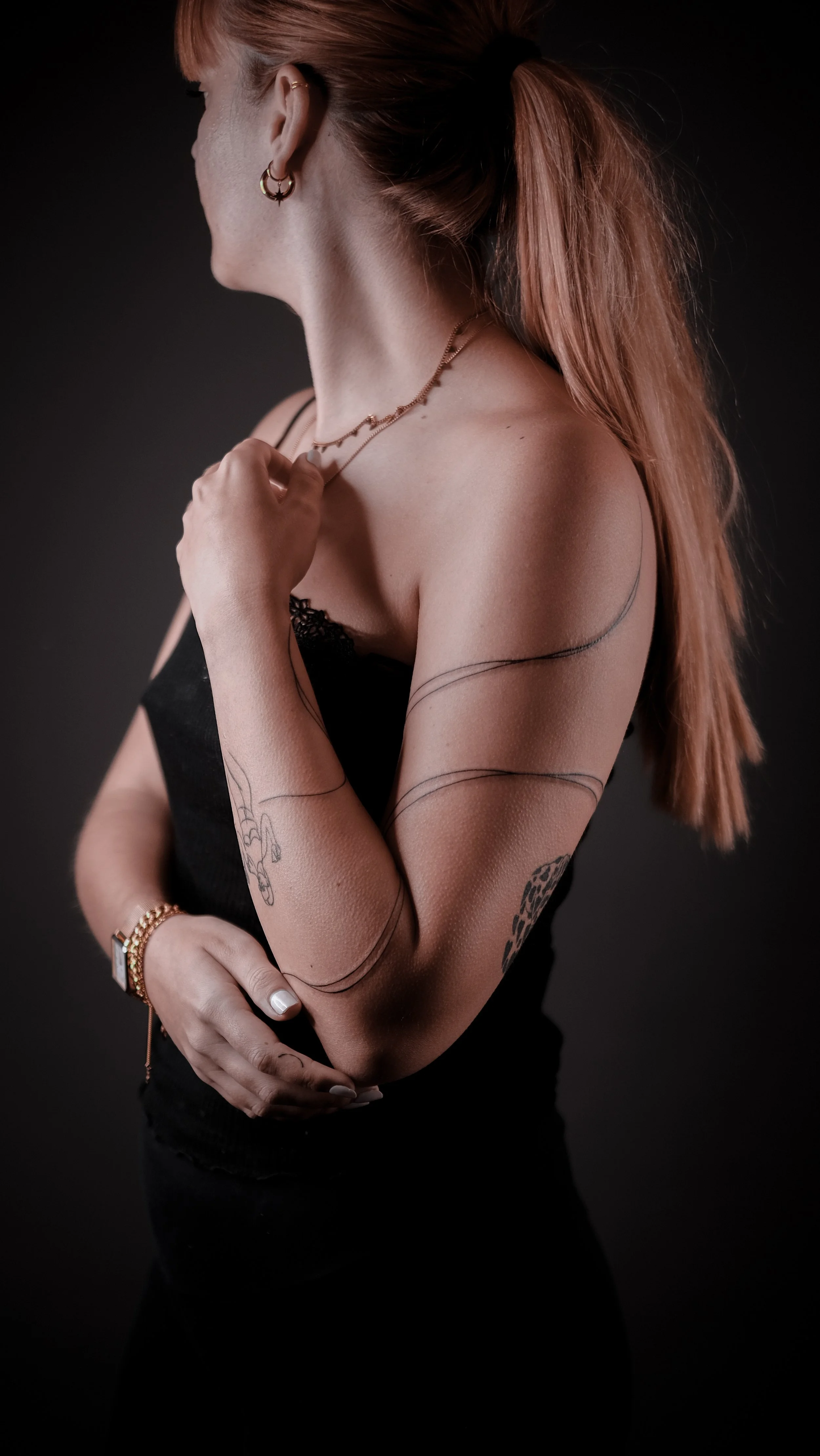 Woman with tattoos on arms, wearing black strapless top, gold jewelry, standing posed sideways.