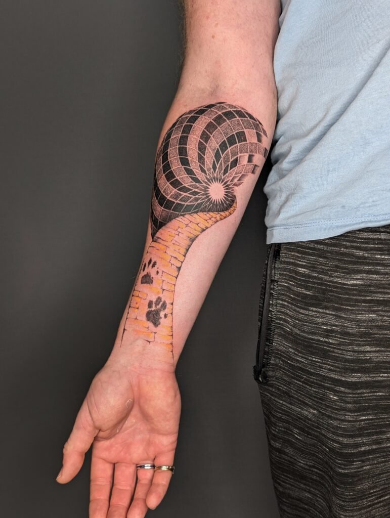 Tattoo of a snake on a person's forearm, with the snake's head near the wrist and its body extending upward. The snake has a brick-like pattern with yellow and orange hues. The head of the snake is in a spiral, creating an optical illusion pattern in black and gray on the upper arm.