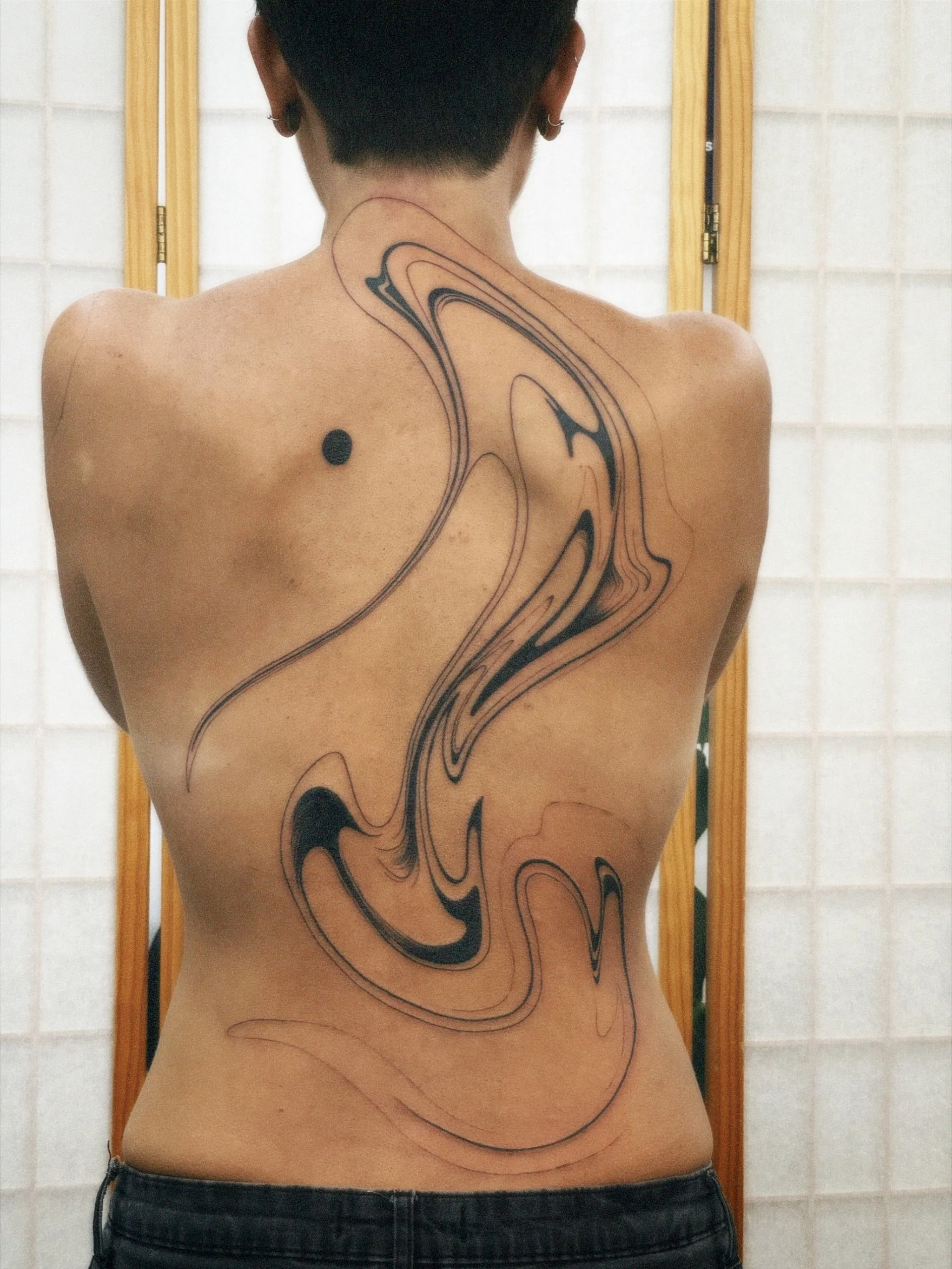 Back view of a person with a large, abstract black and beige marbled tattoo on their upper back and spine.
