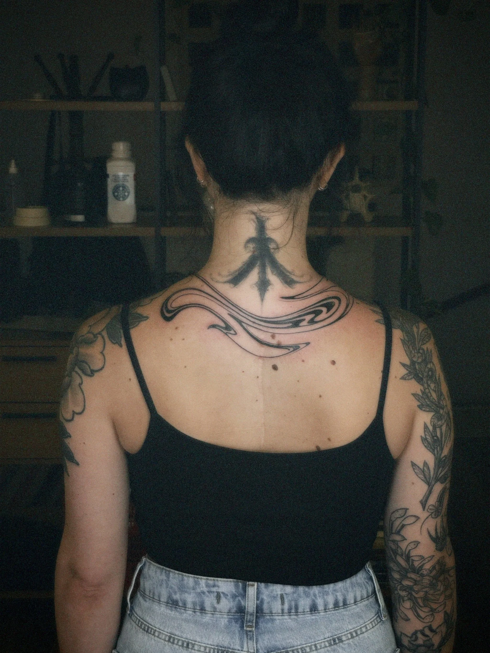 Back of a woman with short black hair, tattoos on her neck, shoulders, and arms, wearing a black tank top and light-colored jeans.
