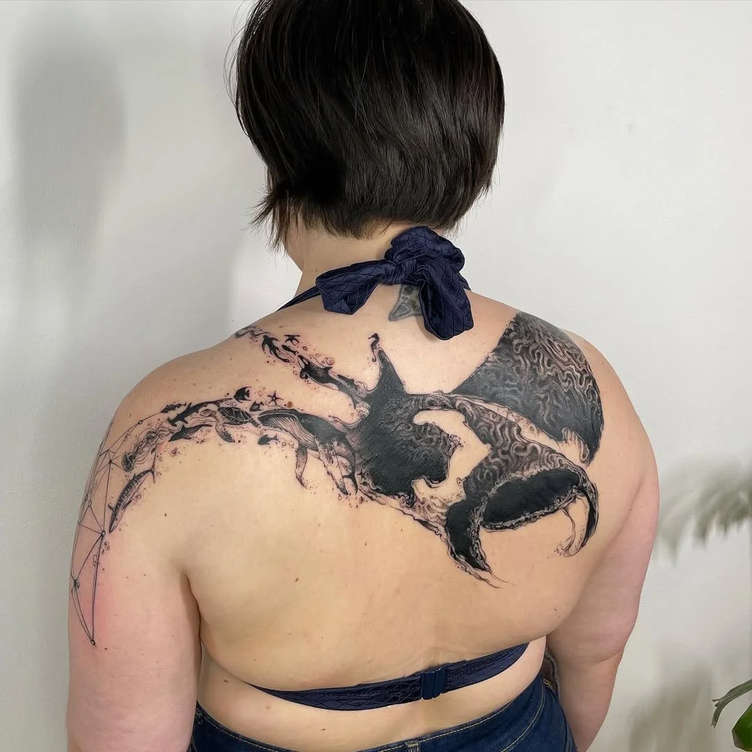 Back view of a person with a large black and white tattoo featuring a dragon design that extends across their upper back and shoulders, with intricate details and symbol-like patterns on the left side. The person has short hair and is wearing a strap