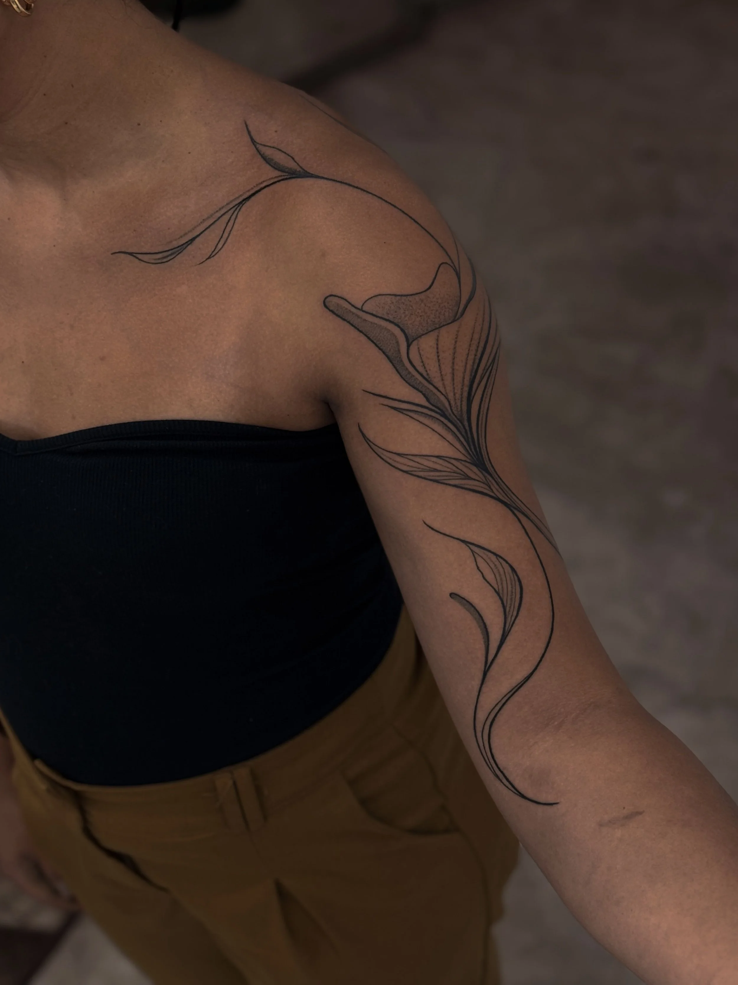 A person's upper arm and shoulder with a black line art floral tattoo extending from the shoulder to the forearm.