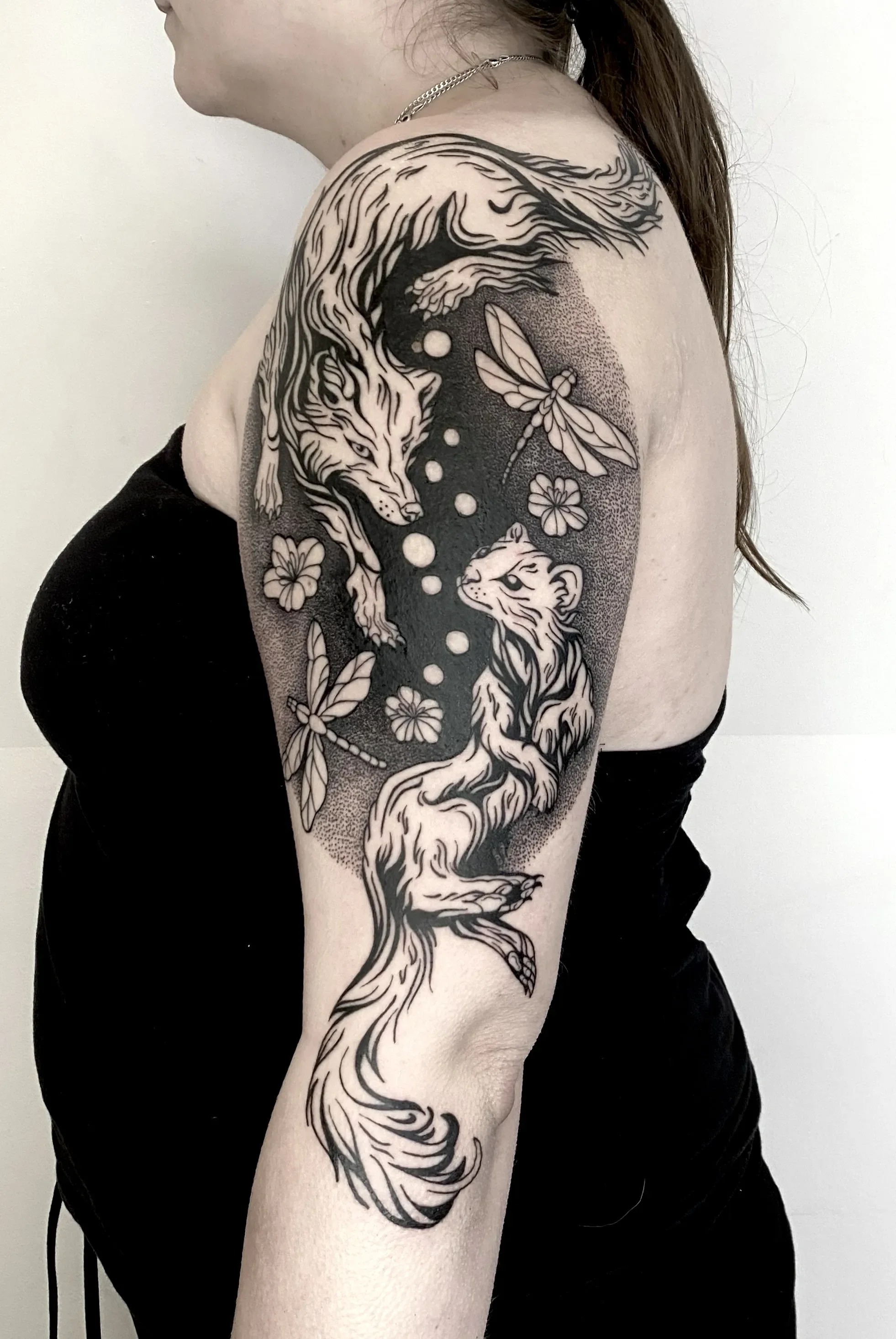 Black and white tattoo of a wolf and a fox on woman's upper arm with flowers and dragonflies.