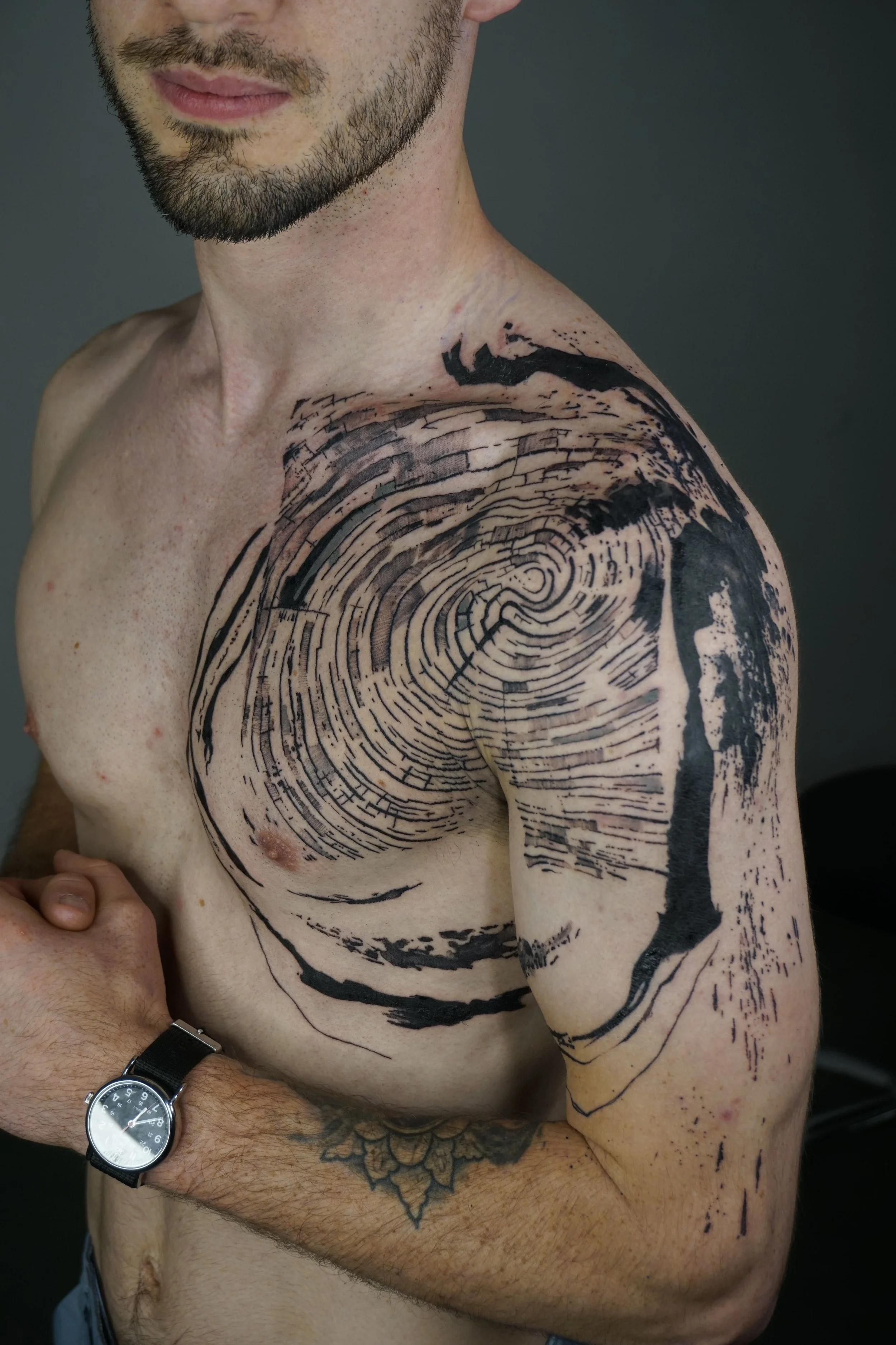 A shirtless man with a beard and short hair, wearing a black watch, shows a large, abstract black tattoo of tree rings and branches on his right shoulder and upper arm.