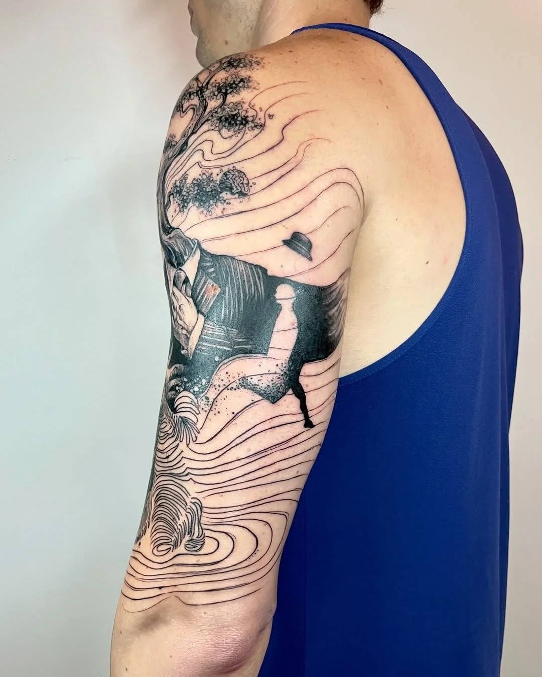 Arm with intricate black tattoo of a tree, waves, and abstract design elements on a person's arm wearing a blue tank top.