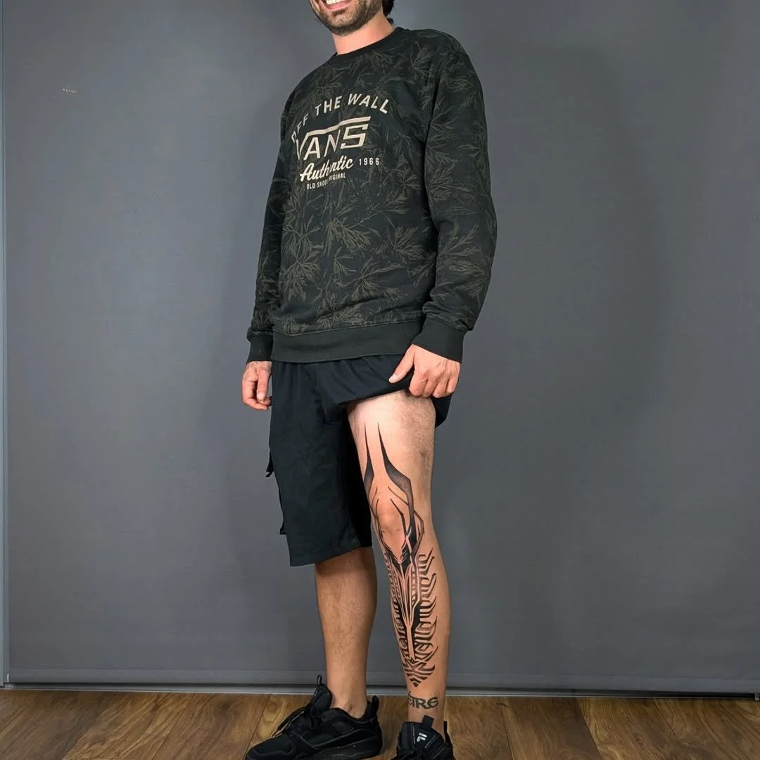 A man standing against a plain wall, wearing a black sweatshirt with white text and graphics, black shorts, and black sneakers. His right leg features a large, detailed tribal-style tattoo extending from his upper thigh to his lower shin.