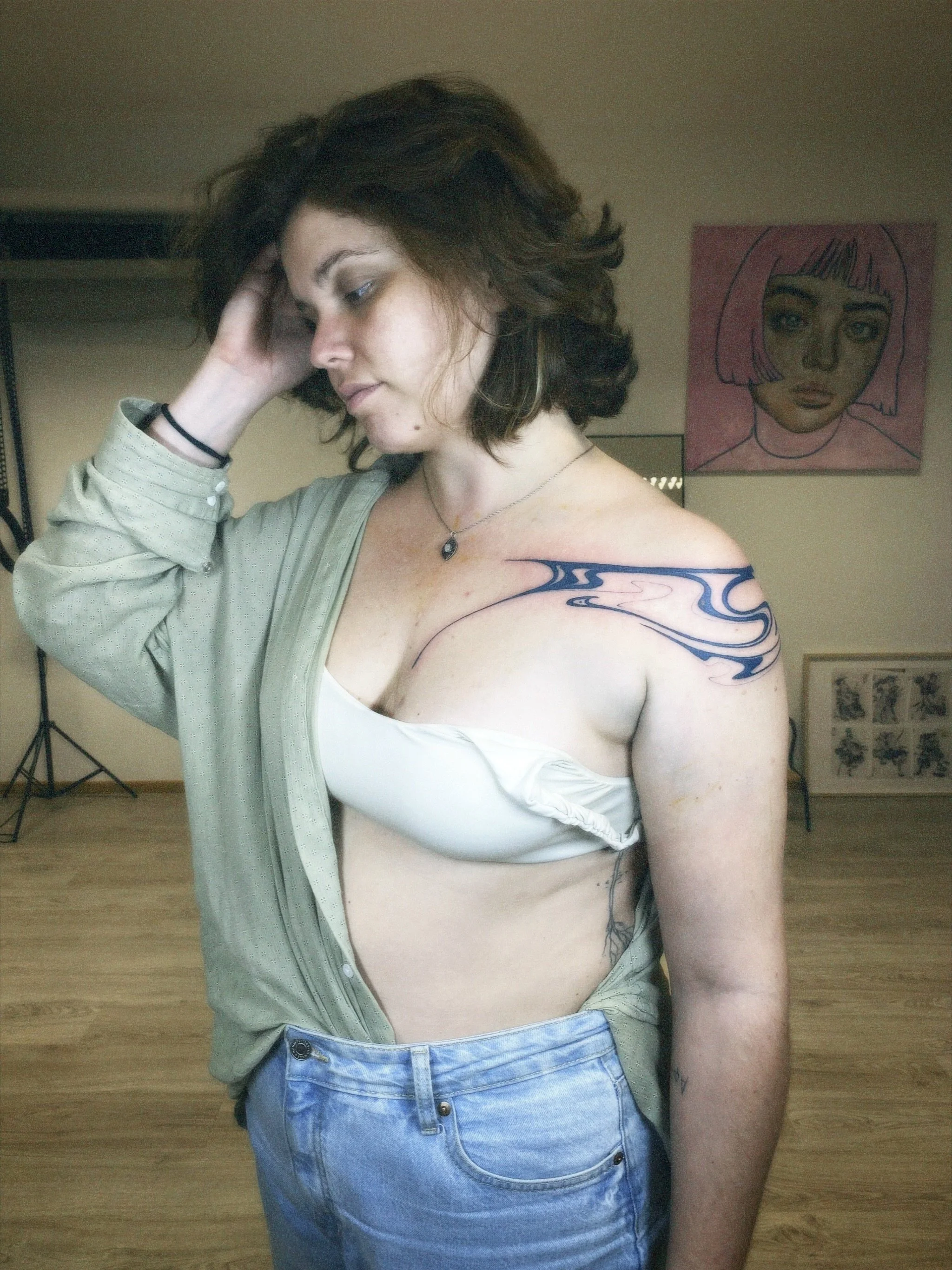 A woman with shoulder-length dark curly hair and tattoos on her left arm and chest, posing indoors with her head tilted and one hand touching her hair. She is wearing a khaki shirt partially unbuttoned, a white slip underneath, and light blue jeans. 