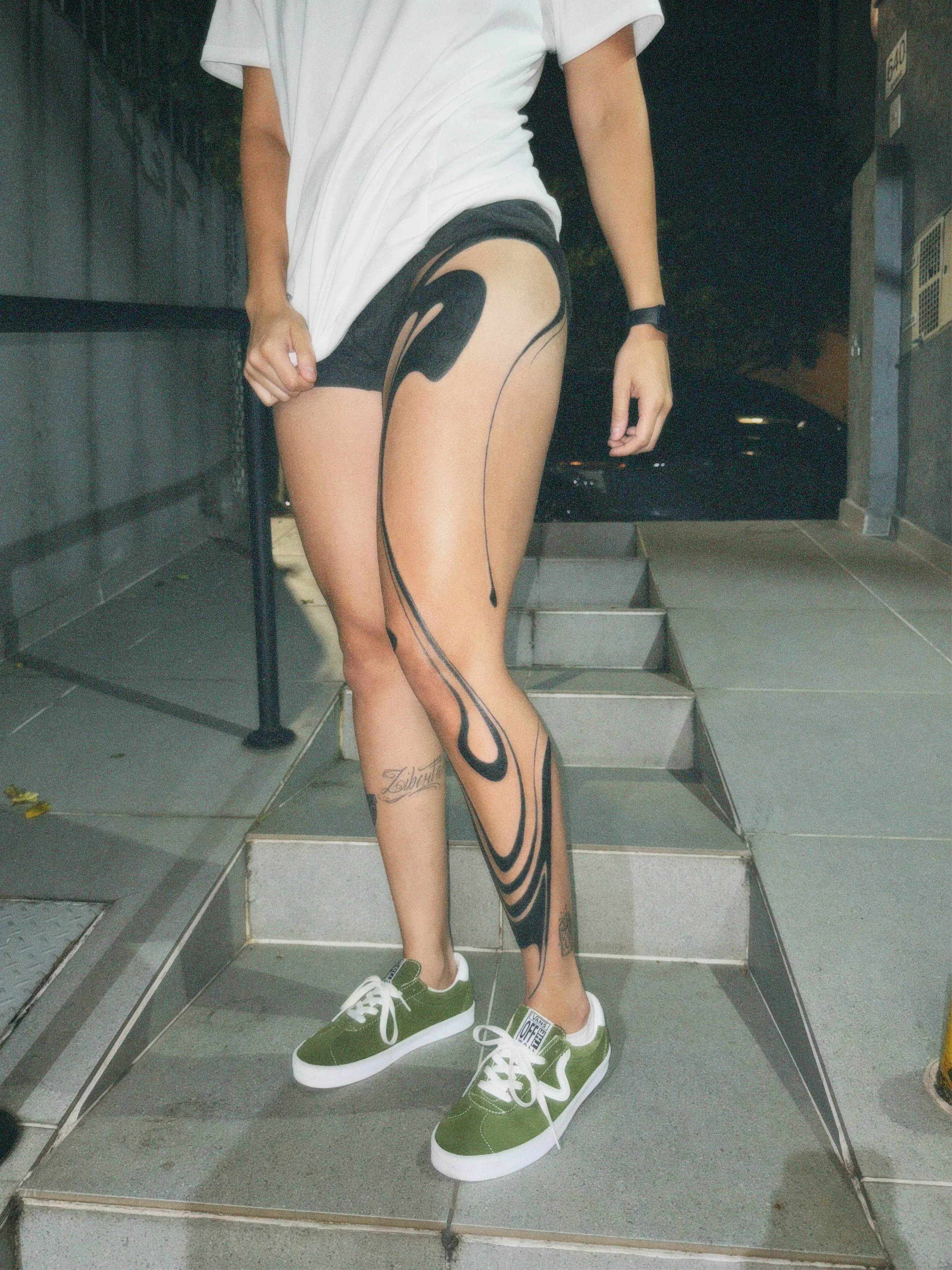 Person wearing an oversized white t-shirt, black shorts, green sneakers, and black tattoo leggings standing on tiled stairs at night.