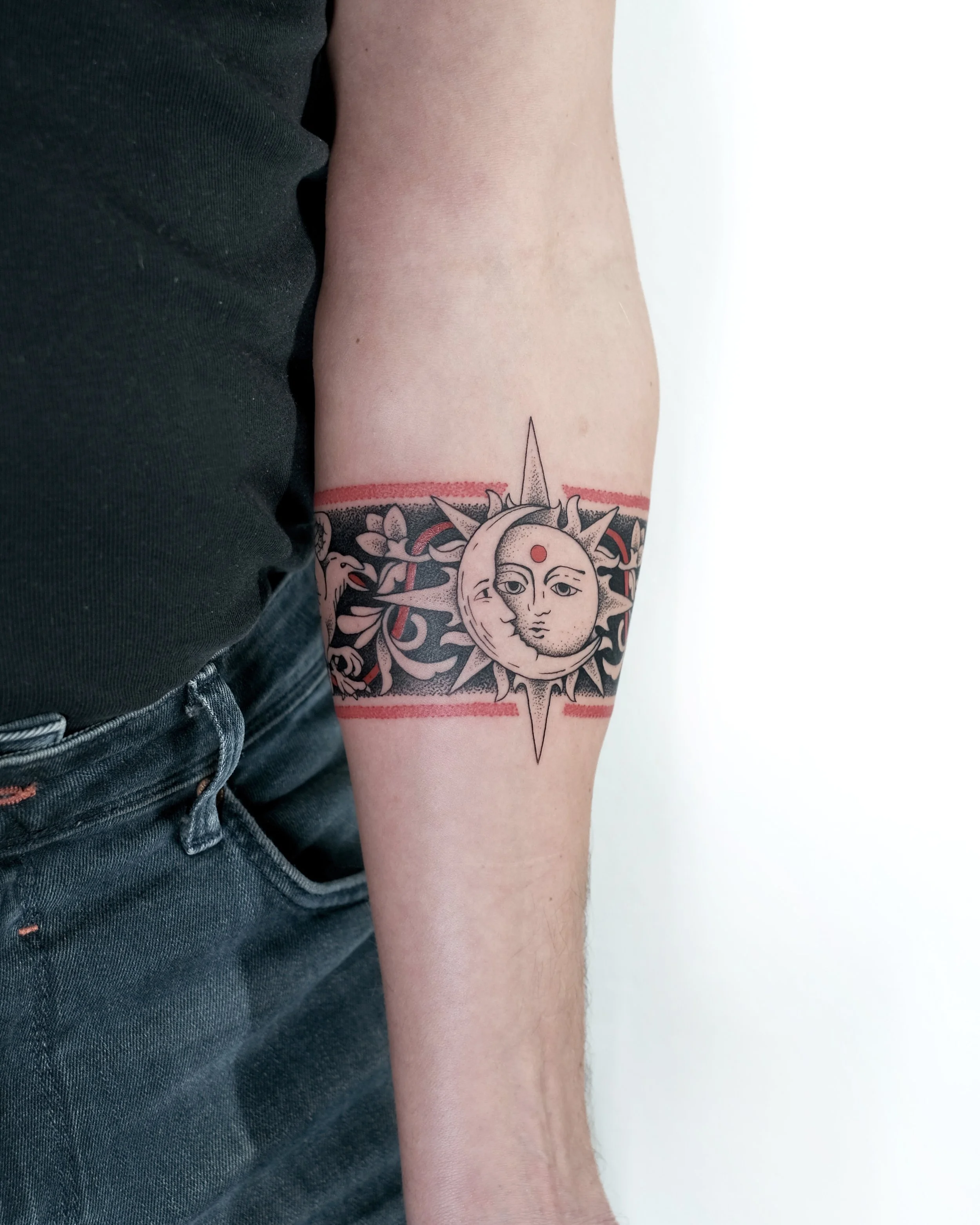Tattoo of a sun and moon face hybrid with a red dot on the forehead, surrounded by decorative black and red floral designs, on a person's upper arm.