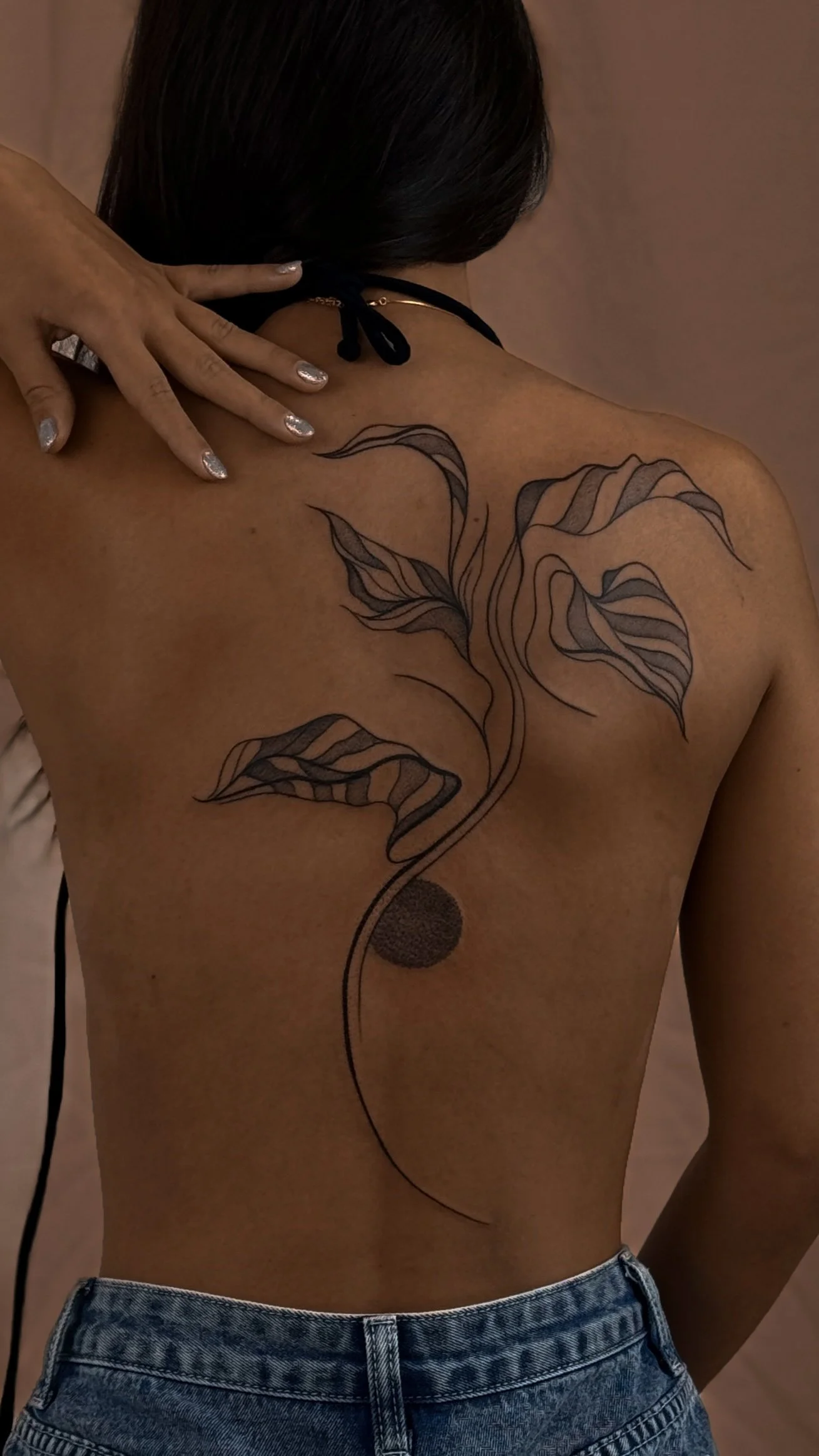 A person with dark hair and glittery nails shows their back with a black abstract floral tattoo and a circle tattoo near the waist, wearing jeans and a black strap top.