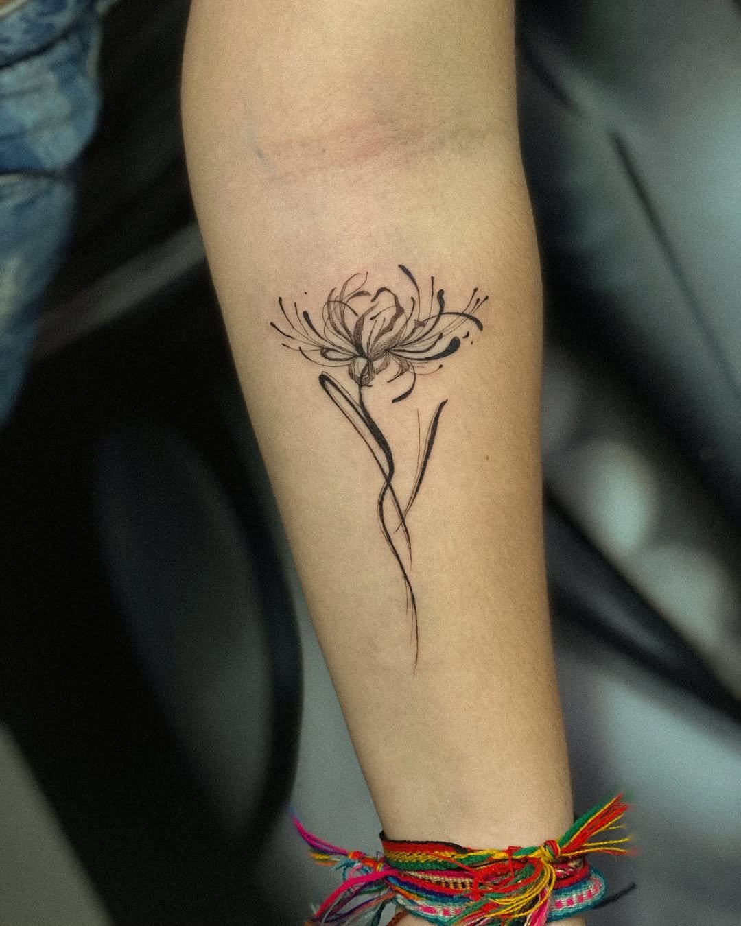 A black ink floral tattoo of a delicate flower with flowing lines on a person's forearm, with colorful string bracelets around the wrist.