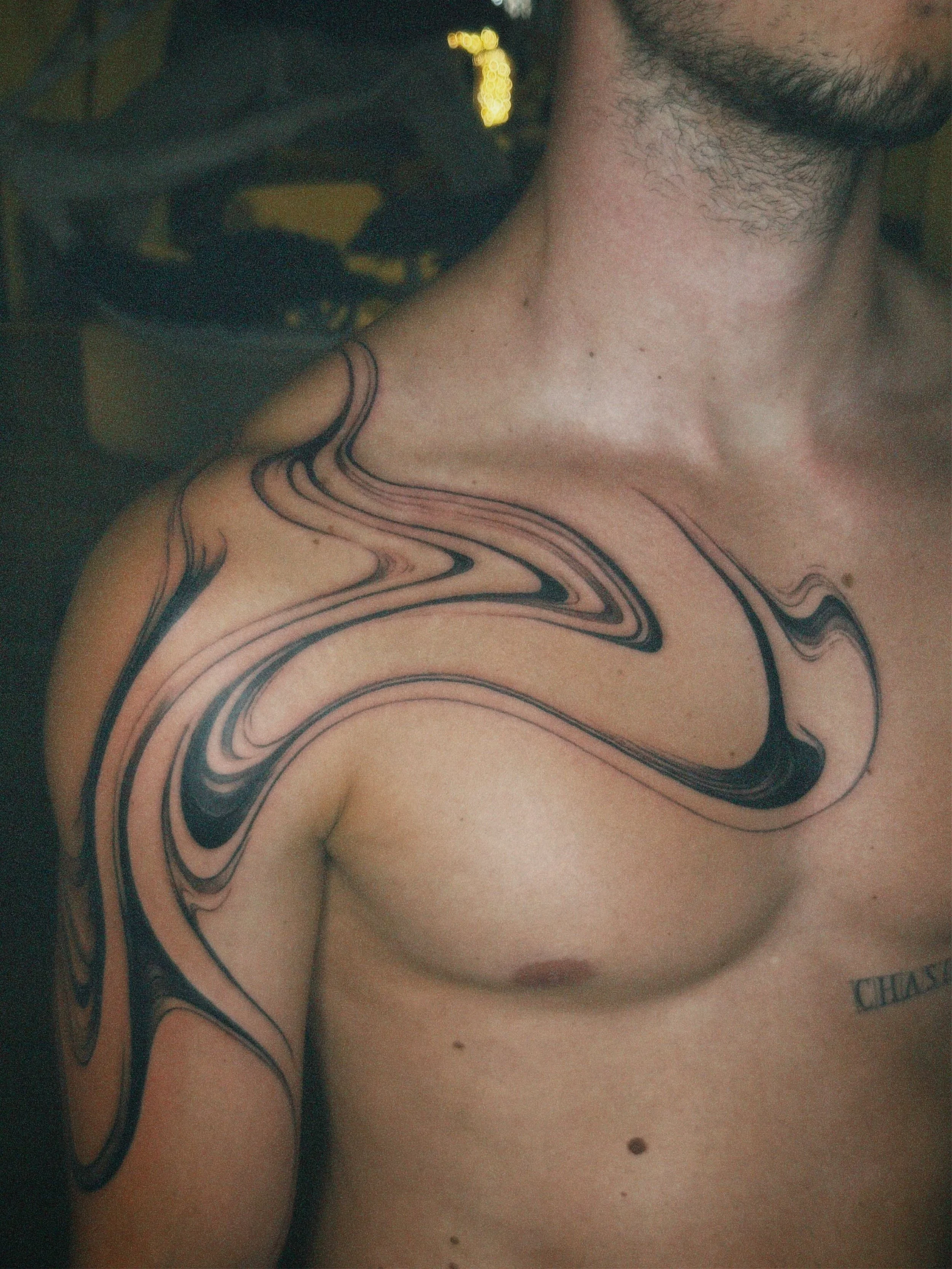 Close-up of a man's shoulder and upper chest with a black and pink abstract swirl tattoo, and the word 'CHASE' tattooed on his chest.