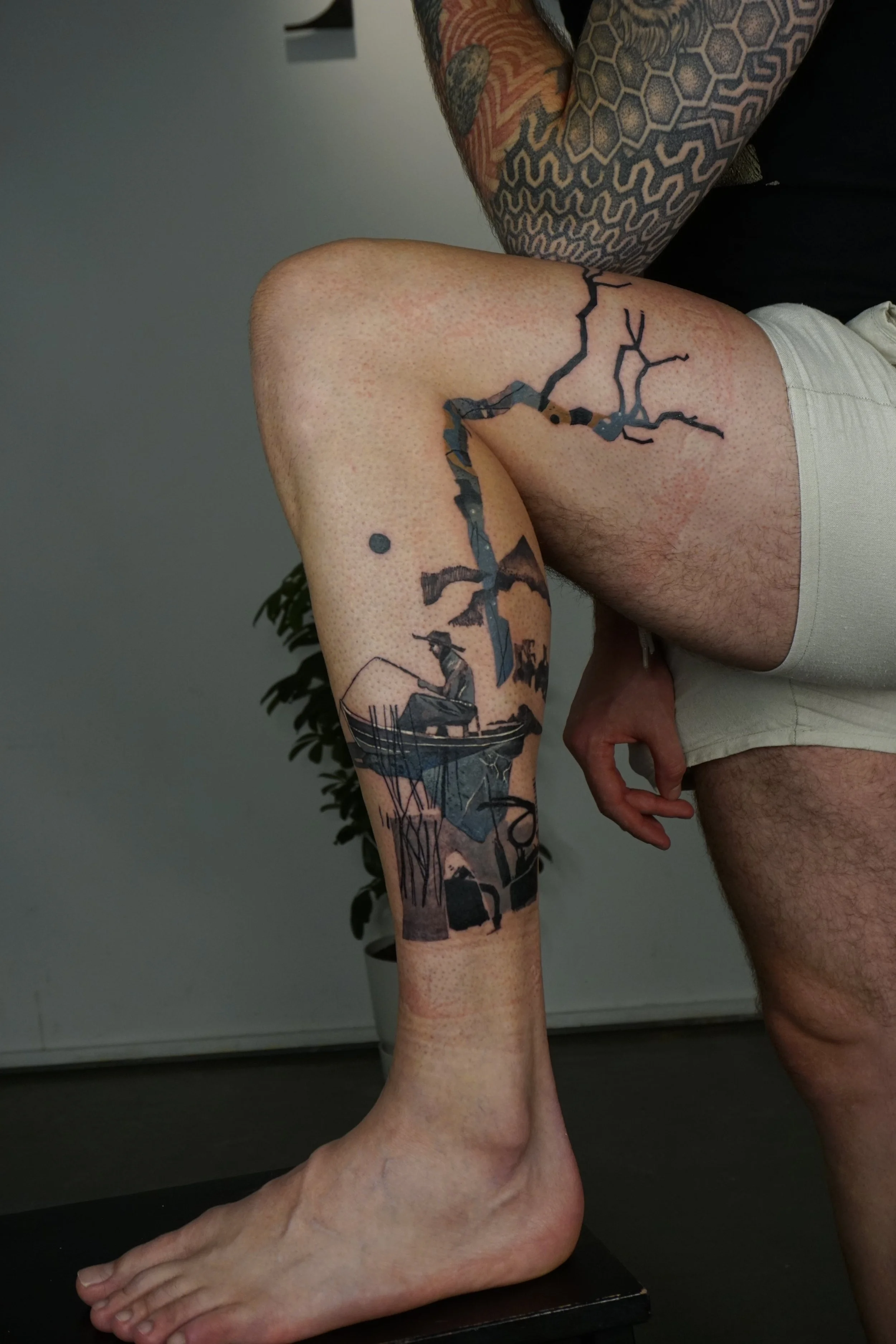 A tattoo of a landscape scene with a boat on water and a large tree, located on the person's leg. The person is sitting with one leg bent, wearing beige shorts, with a tattooed arm resting on their knee. The background shows a plant and a plain wall.