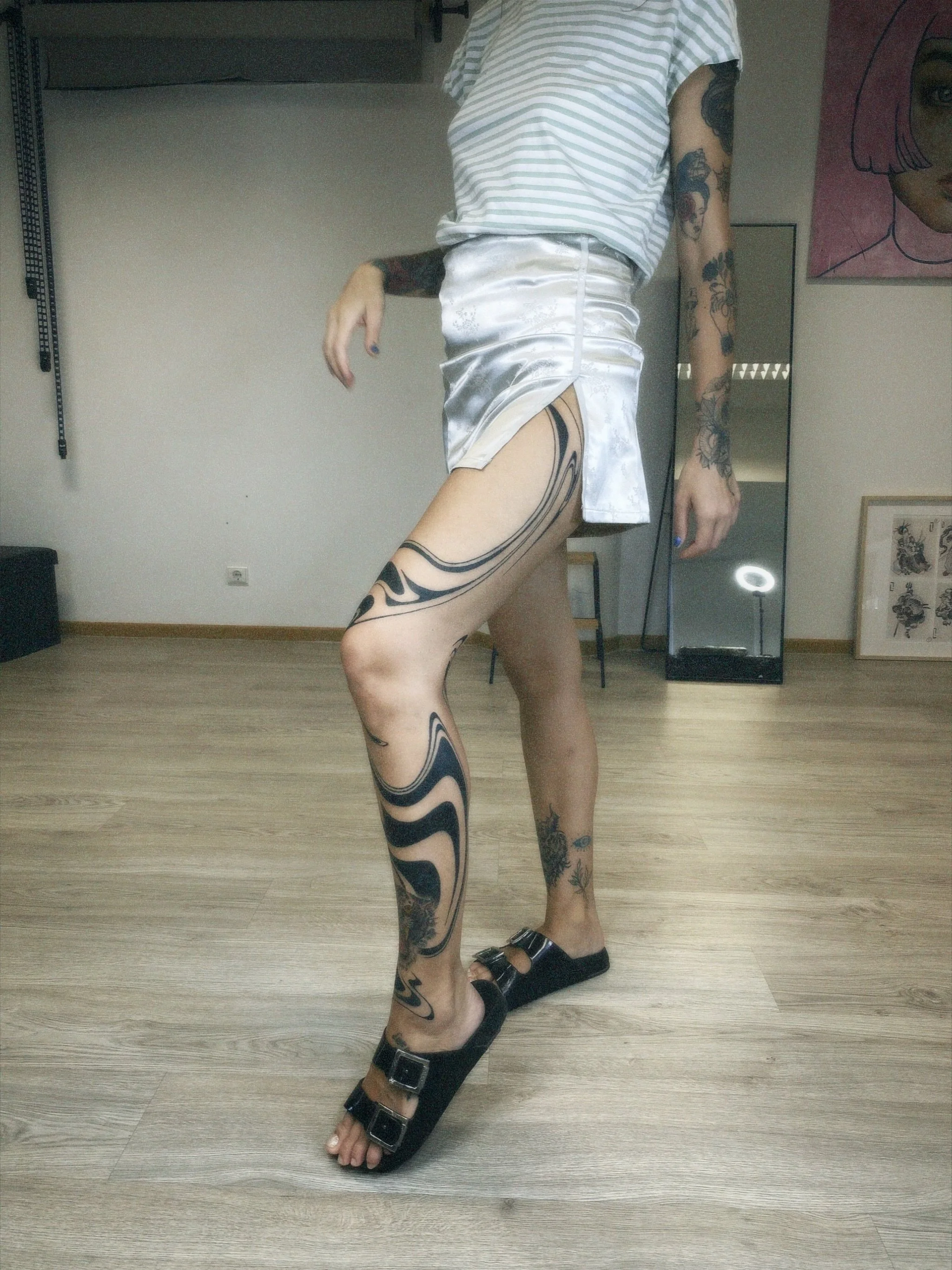 A person standing in a room with tattoos on their arms and legs, wearing a striped top, silky white shorts, and black sandals, with a full-length mirror and framed artwork in the background.