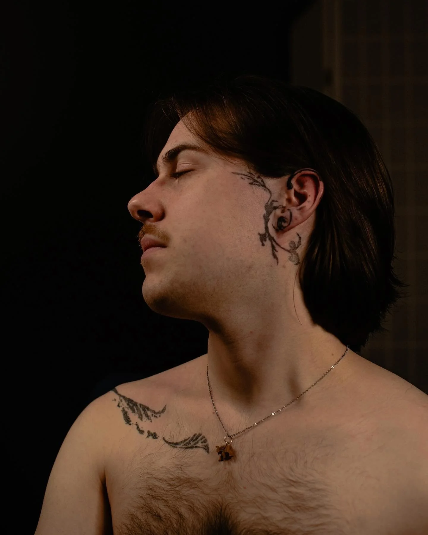 Person with tattoos and necklace, eyes closed, against dark background.