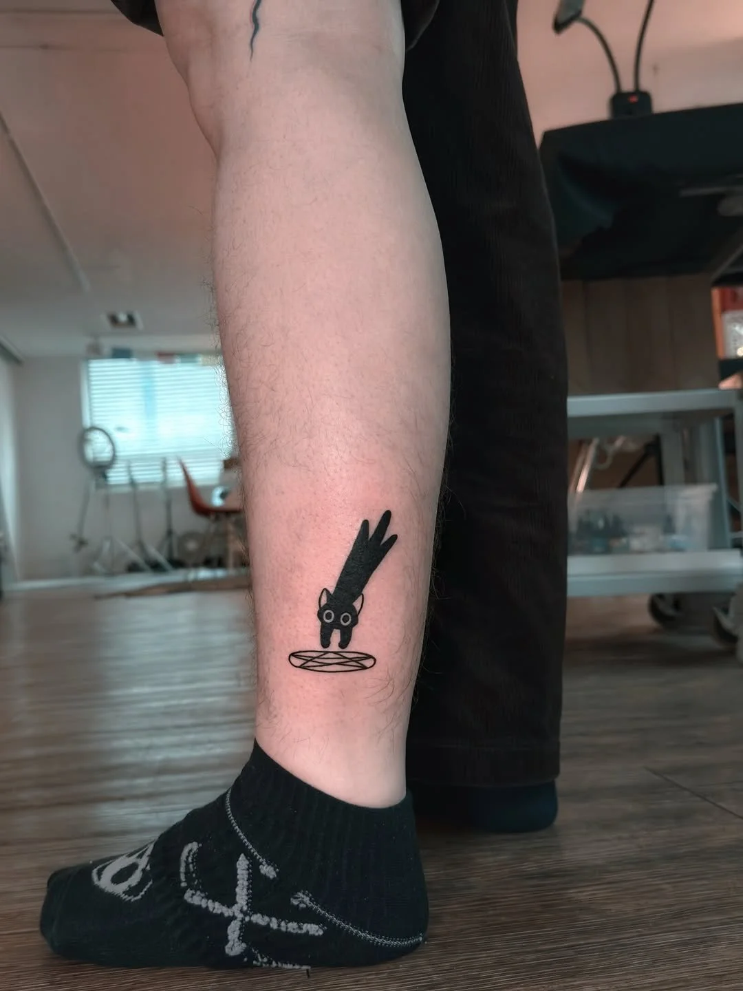 Tattoo of a black cat jumping over a stylized plate or dish on a person's lower leg.