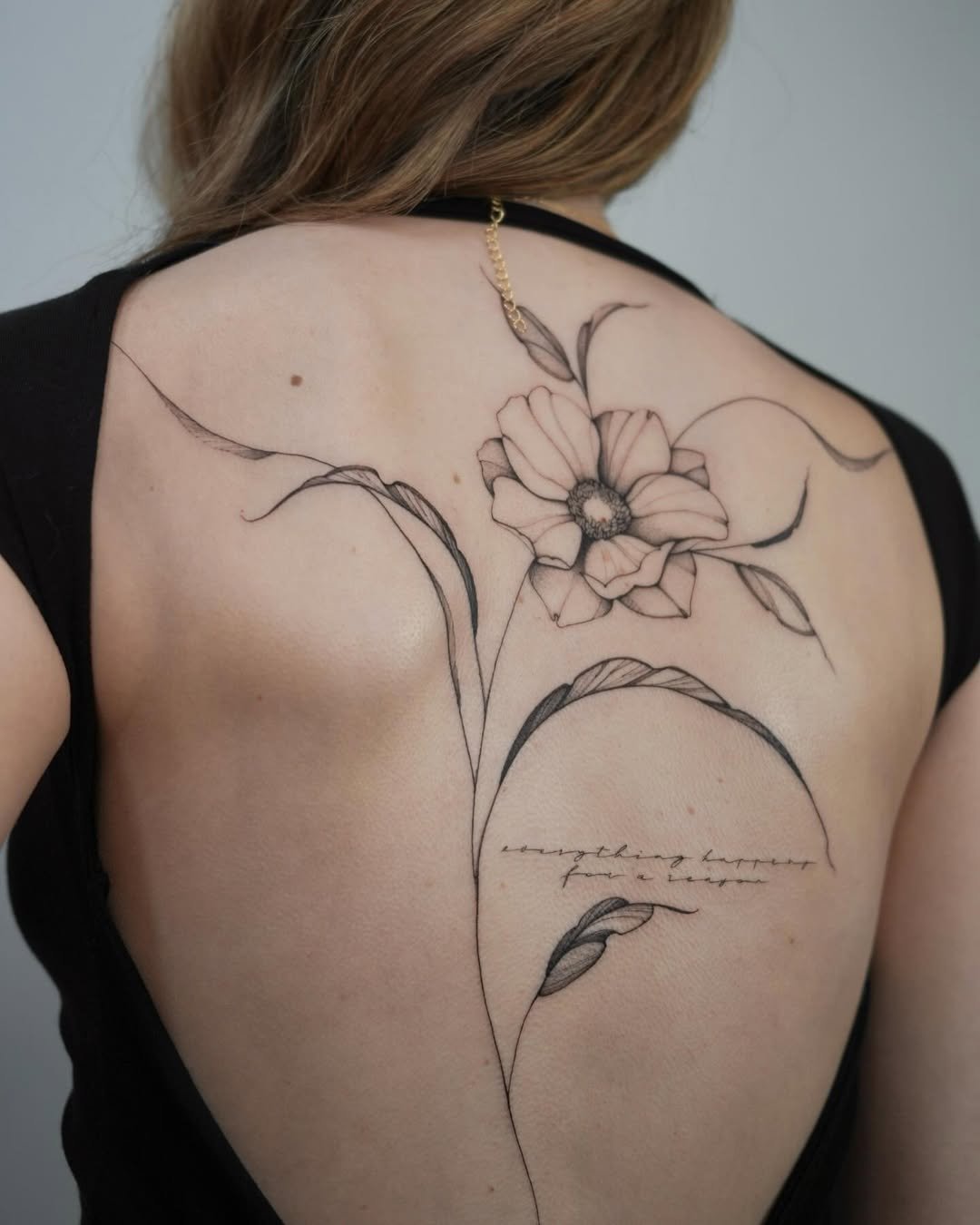 A woman's back with a large, delicate floral tattoo featuring a flower and leaves, along with a quote in cursive writing.