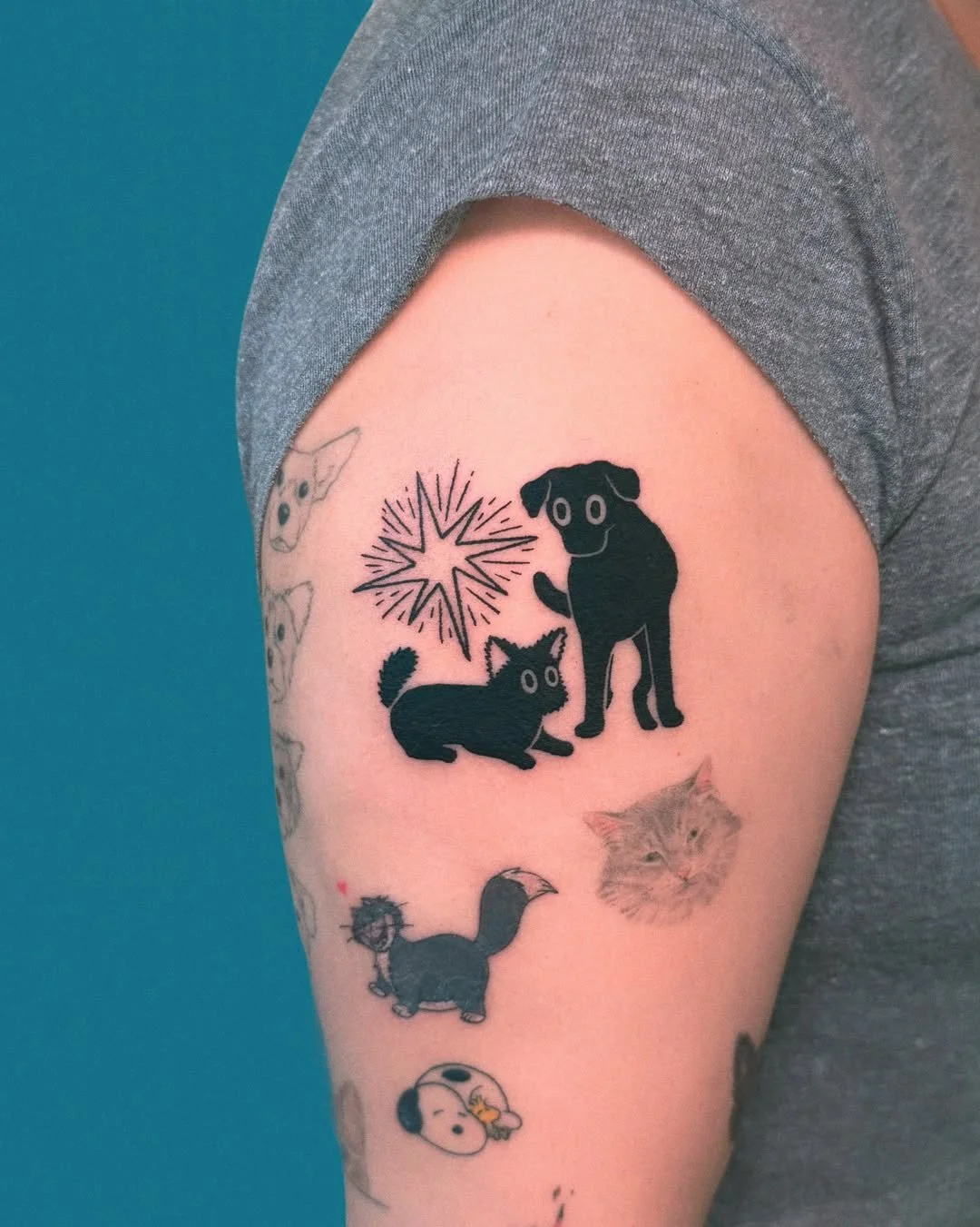 Tattoo of two black dogs with a starburst design above on a person's upper arm, surrounded by additional smaller tattoos of animals, including a cat, a bird, and Peanuts character Snoopy.