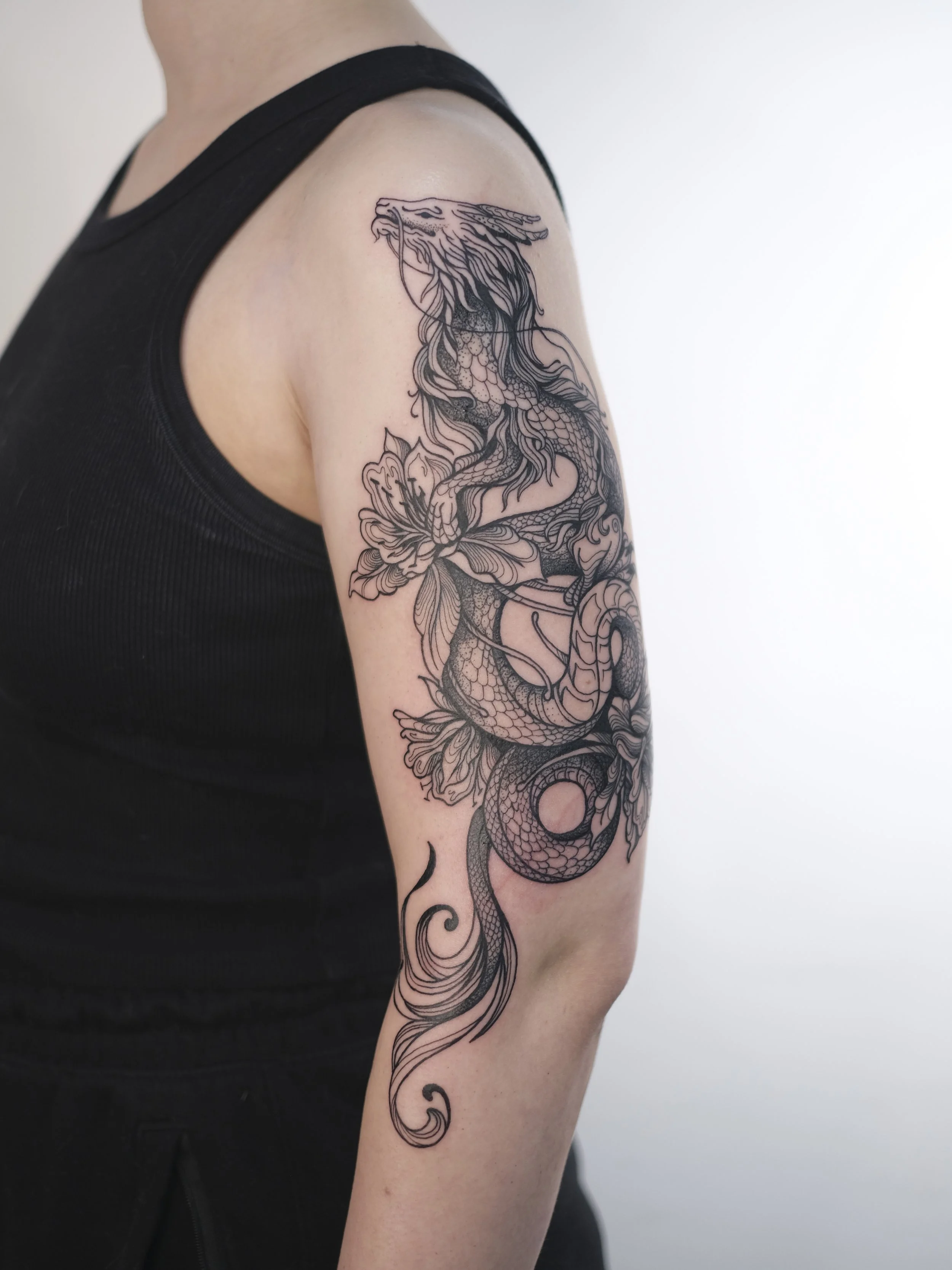 Black outline tattoo of a dragon on a person's upper arm, with intricate details of scales and flowing mane.
